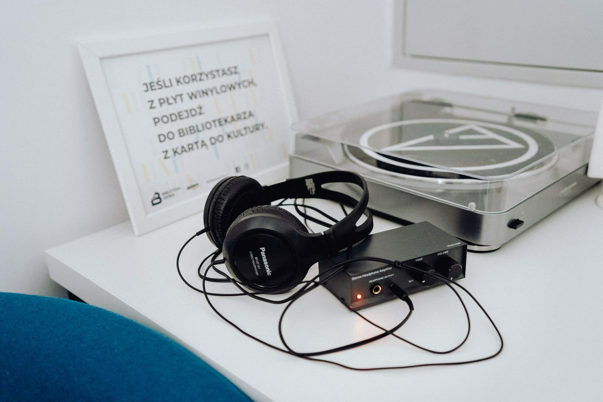 On a white shelf stands a turntable with black headphones and a small black audio device connected to it. Behind the headphones a framed plaque with Polish text, suggesting a photo-report of the event. Part of a turquoise chair is visible in the foreground.  