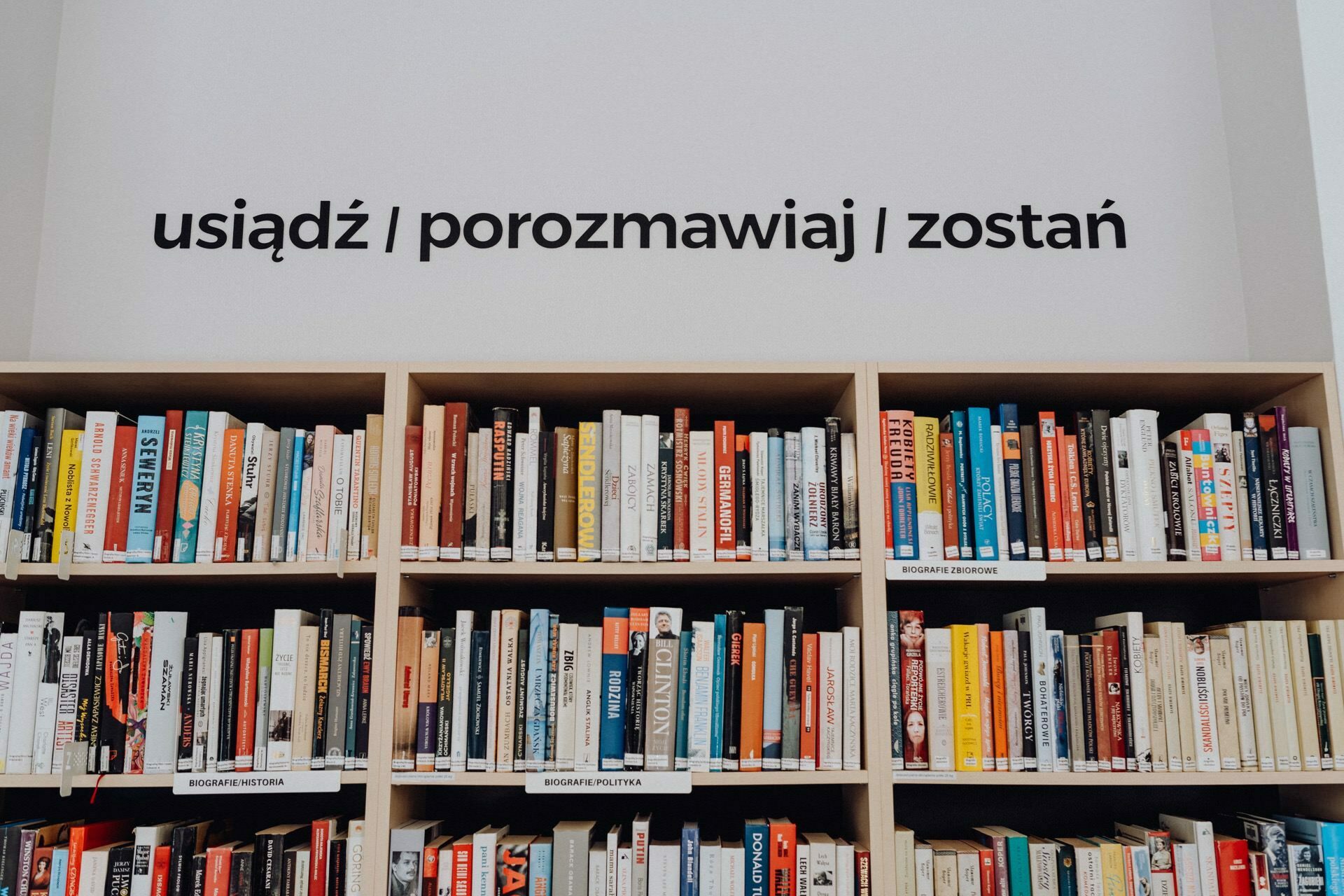 A neatly arranged bookshelf filled with various books. Above the bookshelf hangs a message in Polish that reads "sit | dominate | stay," which means "sit | talk | stay." The calm atmosphere is ideal for readers and could easily be captured by a photographer from Warsaw.  