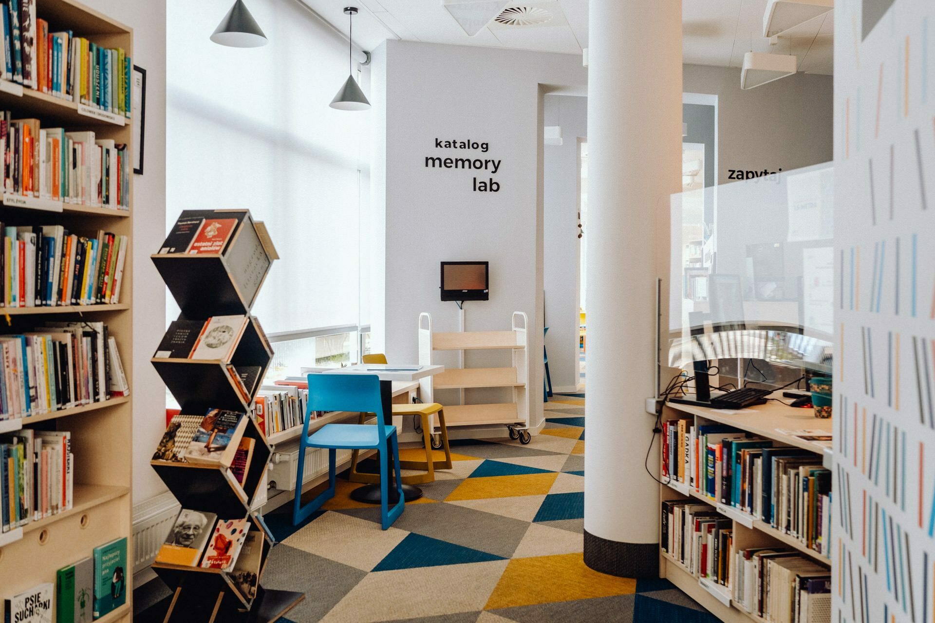 A modern library interior with shelves filled with books, a hexagonal display stand next to colorful chairs and a table, and a computer workstation labeled "catalog memory lab." The room, often photographed by *a photographer from Warsaw*, has large windows to allow natural light to illuminate the space. 