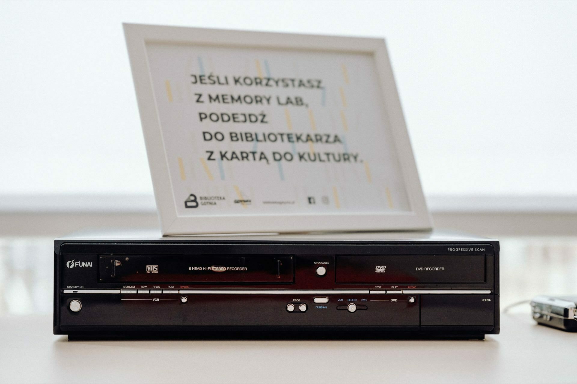 A black retro VHS and DVD player stands on a white surface. Behind it is a framed sign with text in Polish and library icons, perfectly captured by a photographer from Warsaw. The sign is slightly out of focus.  