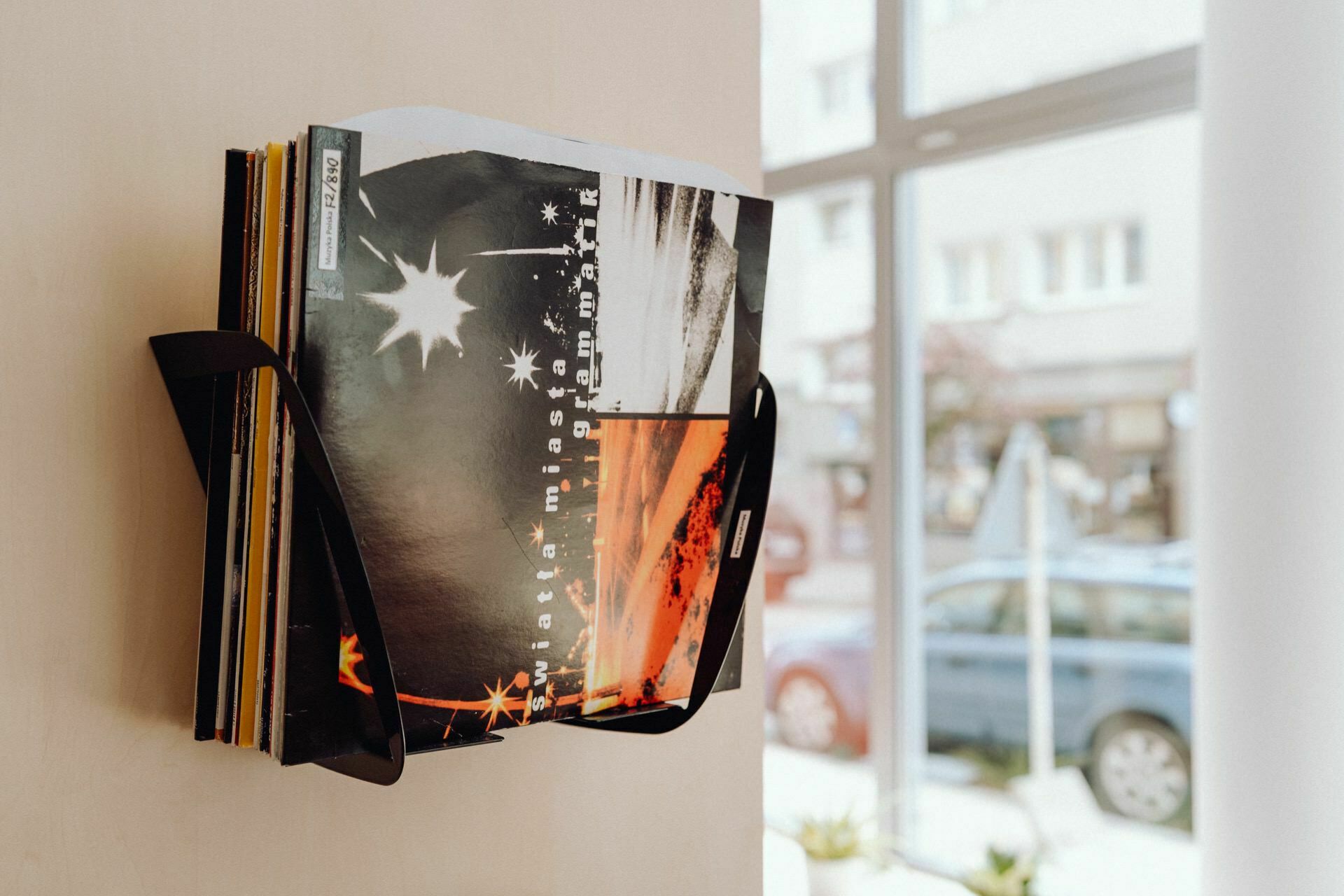 A stack of vinyl records is neatly stored on a black wall rack next to a large window. The discs with colorful album covers have abstract graphics in black, white and orange. Outside the window you can see buildings and a parked car. One can almost imagine *event photographer warszawa* capturing this cozy scene.   

