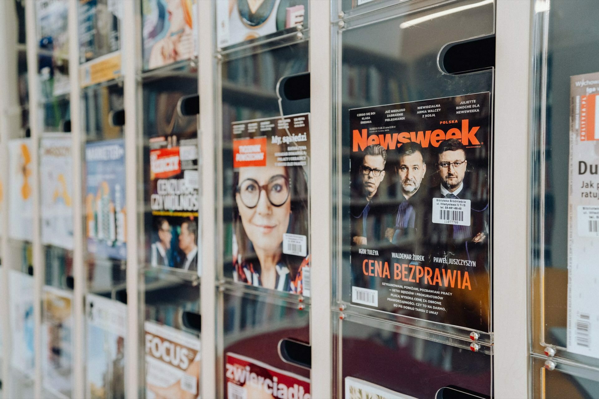 A display shelf filled with various magazines in the store. On the right is a prominent issue of "Newsweek" with three men on the cover. The magazines have colorful covers, including one with a "photo story from the event," and are neatly displayed in clear racks.  