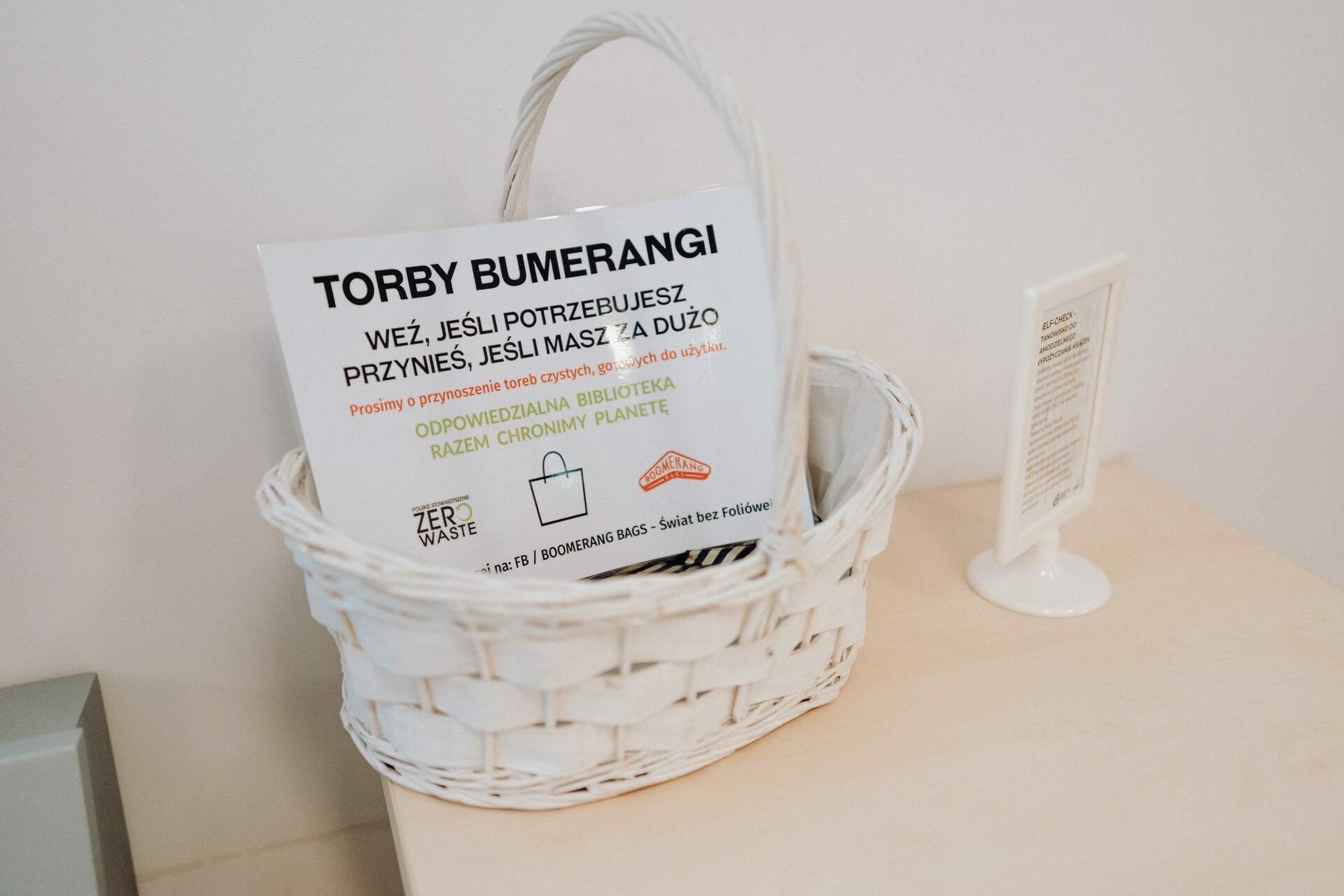 On the table is a white woven basket, and in it are several flyers with Polish text entitled "Boomerang Bags" and mention of "Zero Waste." Next to the basket is a small white sign on a stand. The background, captured by a photographer from Warsaw, is a plain white wall, which adds to the simplicity of the photo coverage of the event.  