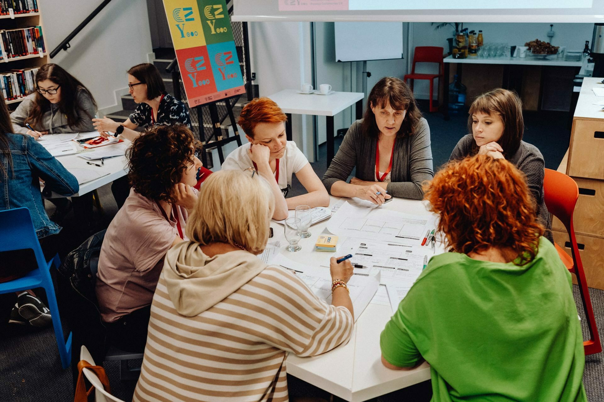 A group of people are sitting at tables in a classroom having a discussion. Papers, notebooks and writing utensils pound on the tables. In the background are shelves of colorful folders and a blackboard. They give the impression of being focused on a common task - perfect for a photo report of the event by a photographer from Warsaw.   