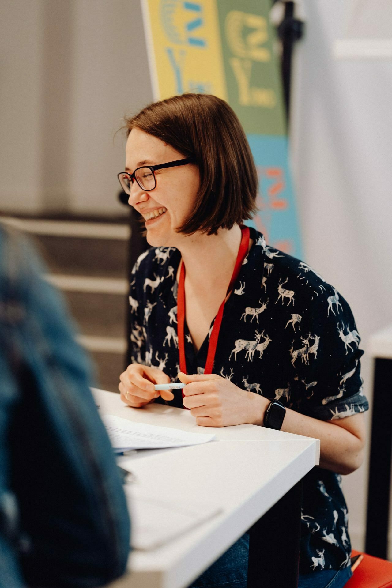 A smiling person wearing glasses sits at the table and carries on a conversation. They wear a dark shirt with a deer print and a red lanyard. In the background you can see a colorful poster and fuzzy figures, perfectly captured by the photographer at the event from Warsaw.  