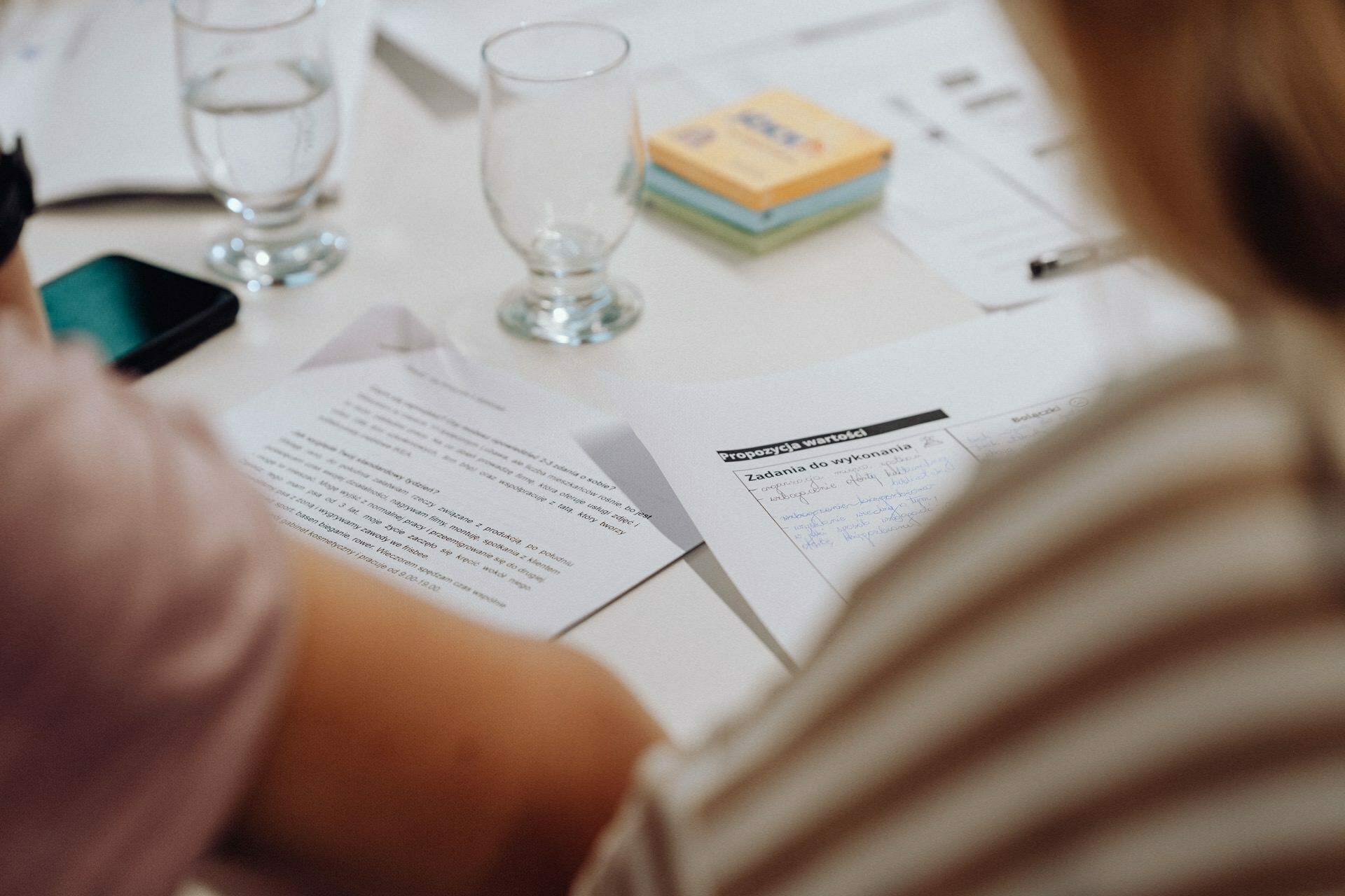 Two people sit at a table engrossed in a discussion or meeting. On the table lie some papers, a smartphone, a bundle of sticky notes and two glasses of water. The focus is on the papers, while the people in the foreground are slightly out of focus - perfect for a photo essay of an event.  