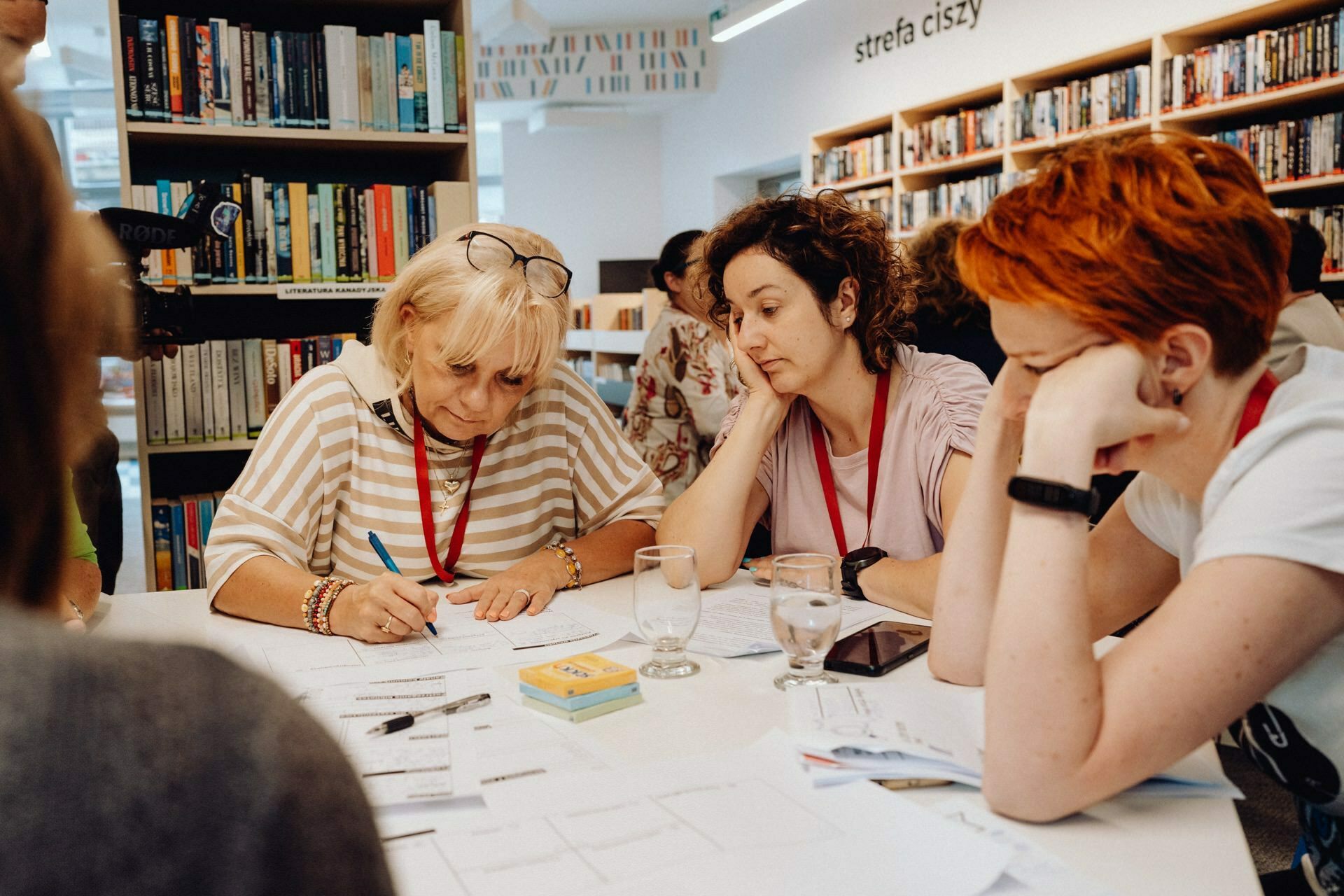 A group of four women sit around a table in a library, immersed in deep discussion or collaborative work. On the table lie papers, notebooks and a glass of water. In the background you can see bookshelves and the sign "zone of silence" (quiet zone). The smooth capture of this moment is thanks to a photographer from Warsaw.   