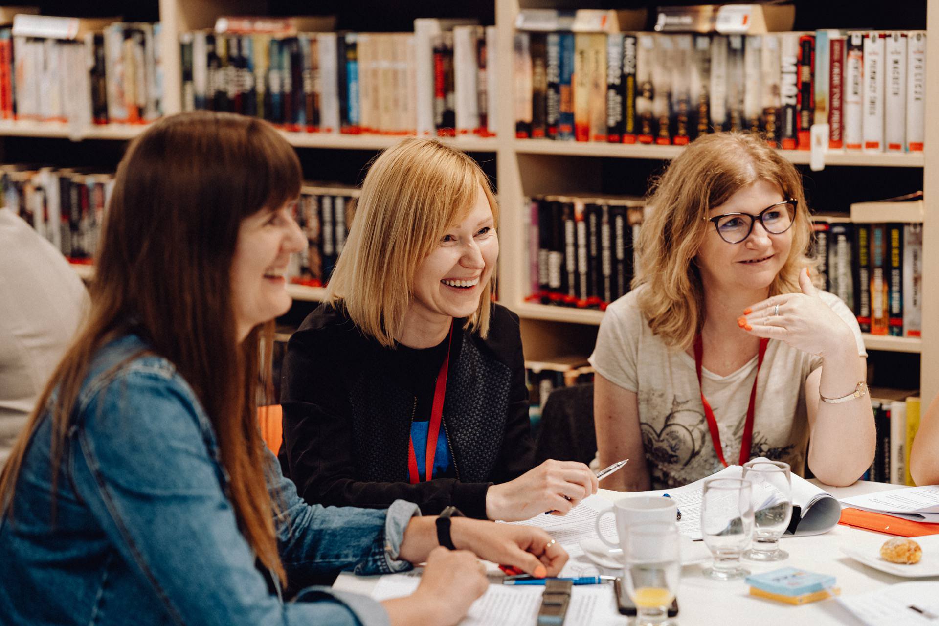 Three people are sitting at a table in the library, engrossed in a lively discussion. They have papers, pens and drinks in front of them. In the background you can see shelves filled with books - a perfect scene that any event photographer Warsaw will capture.  