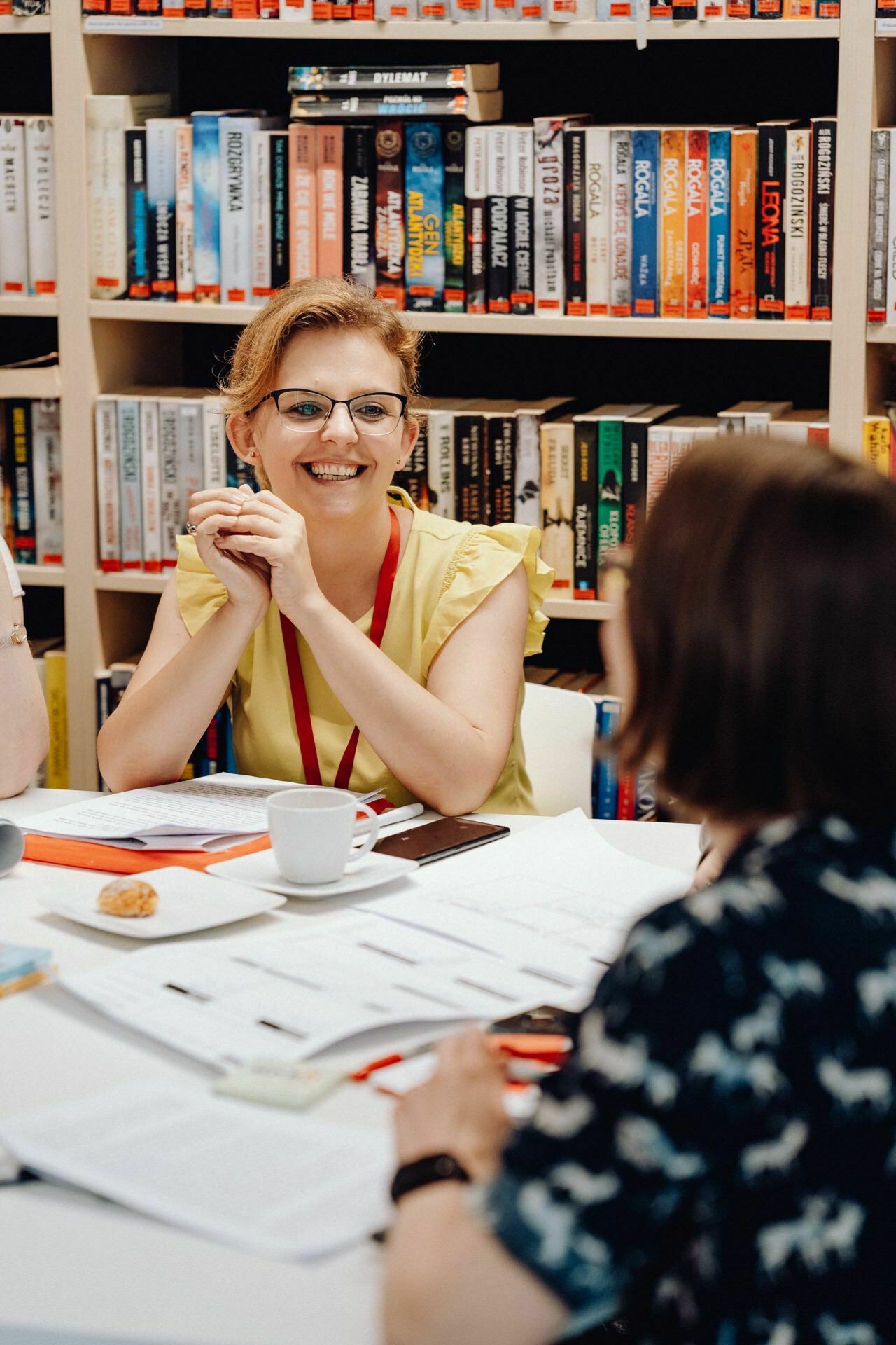 A woman wearing glasses and a yellow shirt smiles, sitting at a table in front of a shelf filled with books. She is having a conversation with another person, with her back to the camera. The scene captures the essence of an animated encounter, perfect for a photographer from Warsaw documenting everyday moments.  