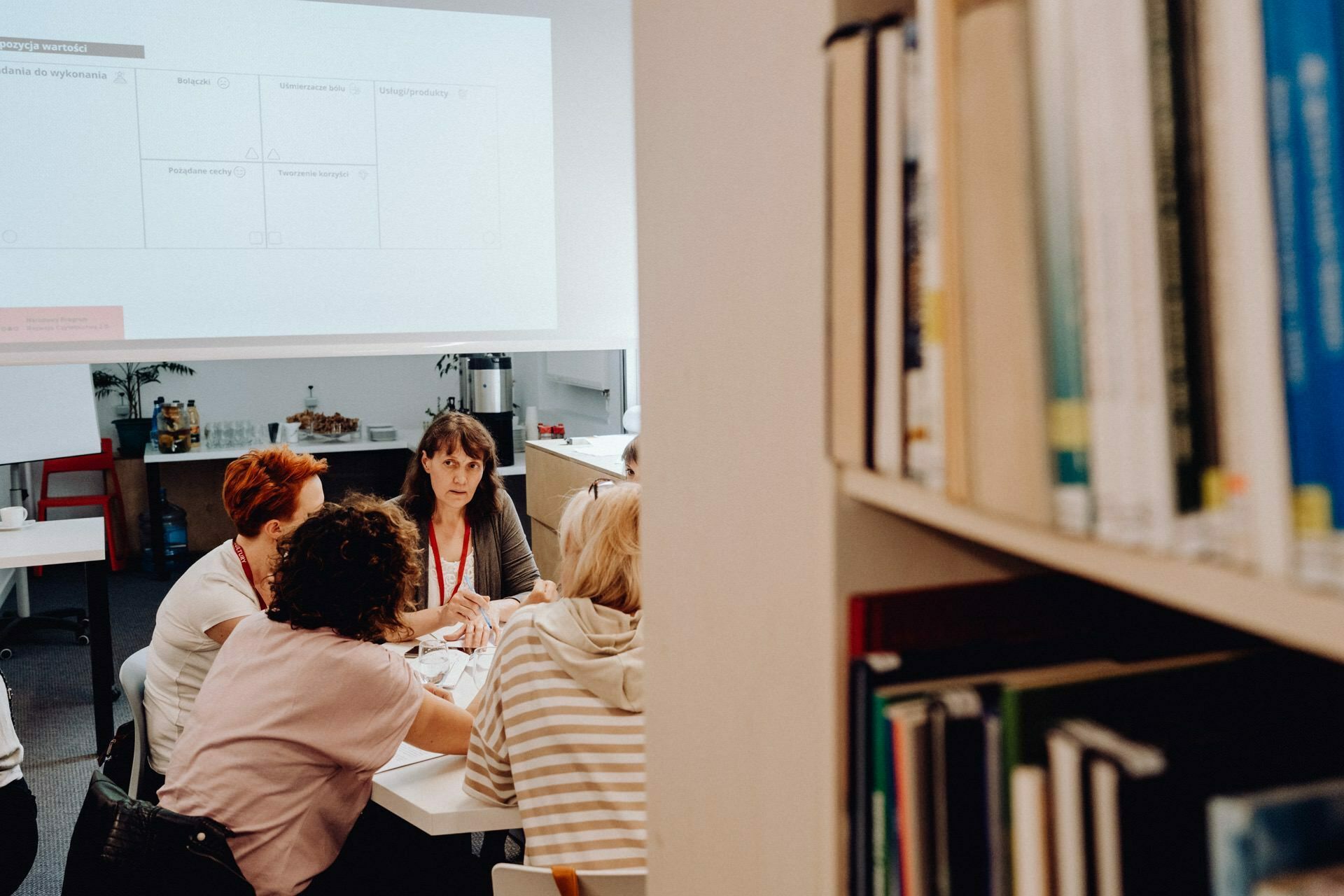 A group of people gathered around a table in a conference room immersed in discussion. In the foreground to the right, shelves of books are visible. In the background you can see a projector screen displaying a chart or diagram, all captured by a skilled event photographer from Warsaw.  