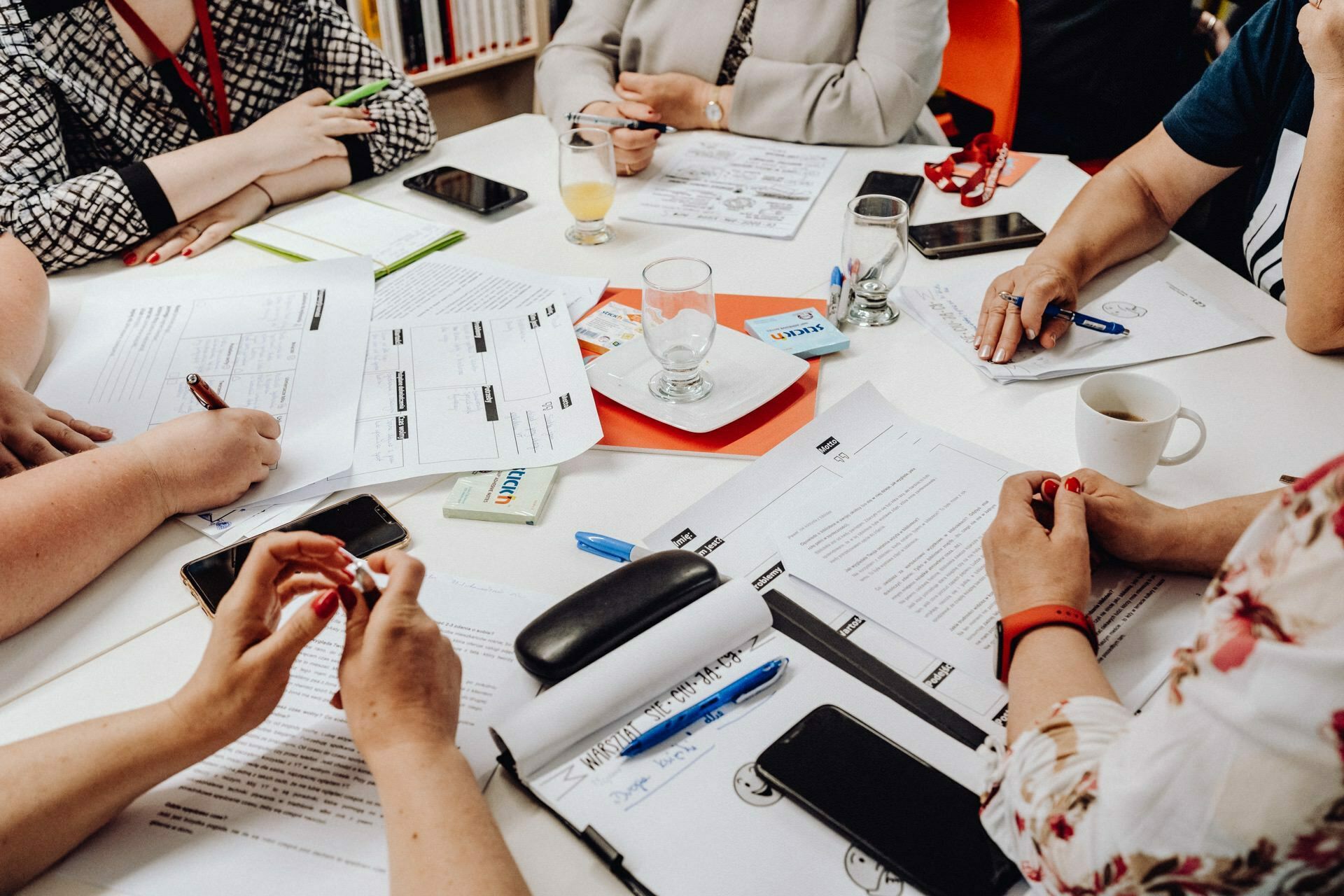 A group of people sit around a white table and collaborate on documents, creating what appears to be a typical photo report of an event. Scattered around the table are various papers, notebooks and electronic devices. One person is drinking coffee and another a glass of orange juice. Only their hands are visible.   