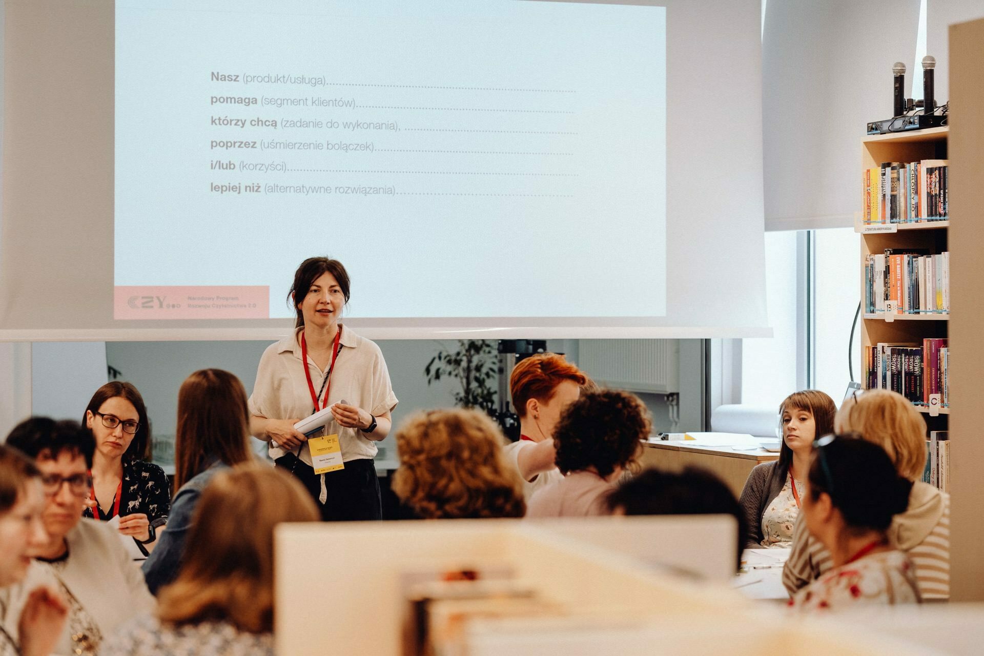 The woman stands in front of a group of seated people and performs in a well-lit room. Behind her, a projection screen displays text in Polish. Bookshelves are visible on the right side of the room. The participants are engaged and appear as if they are participating in a workshop, which was beautifully captured by our event photographer Warsaw.   