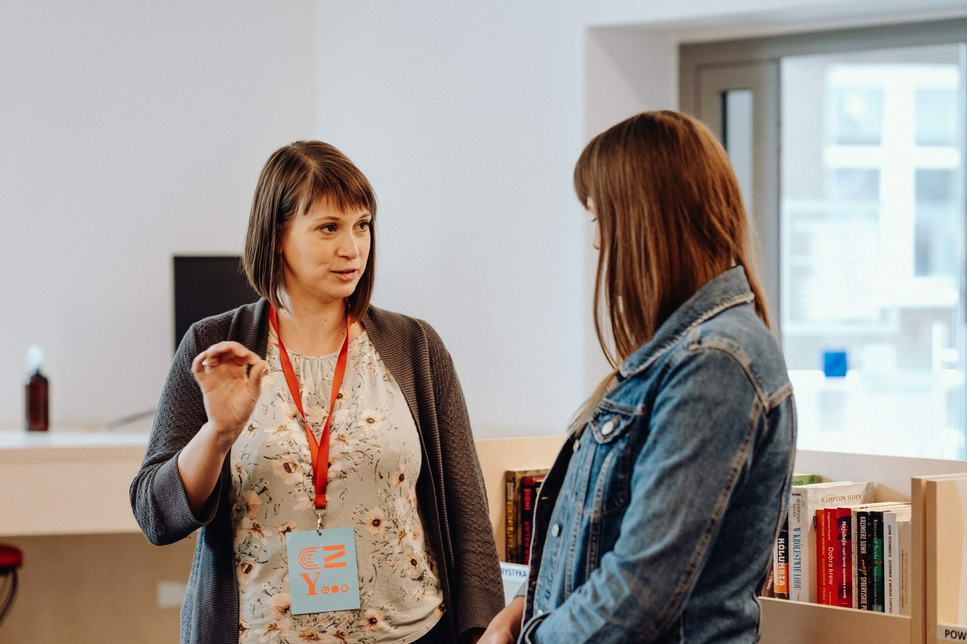 Two women are engaged in conversation in what appears to be a library or bookstore. The woman on the left, wearing a name badge, gestures animatedly. The woman on the right, wearing a denim jacket, listens intently. They are surrounded by shelves of books - the perfect setting for an event photo essay captured by event photographer Warsaw.   