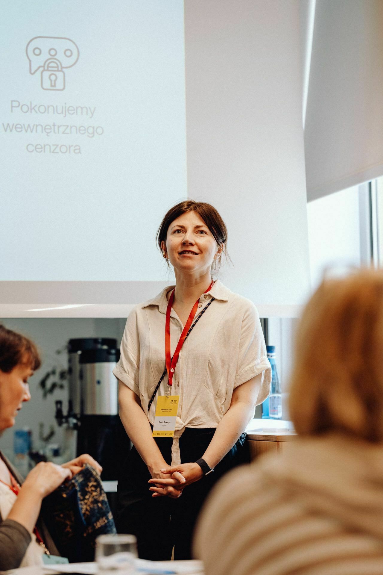 A person stands and speaks in front of a group in a room. They wear a white shirt, a red lanyard and a name badge. A slide behind them displays text and graphics. The text of the slide appears to be in Polish. The other participants sit and listen attentively, as if waiting for the photo-report of the event to begin.    