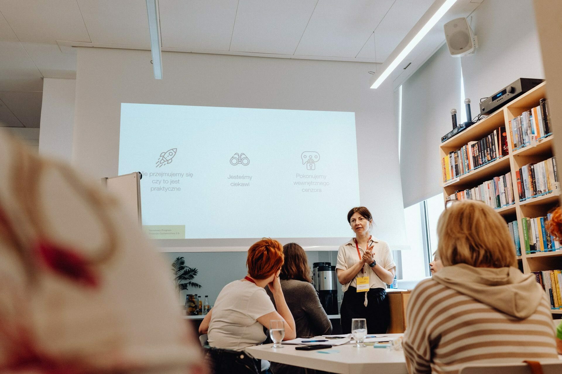 A woman gives a presentation in the room while participants sit at tables. A projector screen behind her displays icons and text. Bookshelves stand along the wall to her right, and the concentration of the participants involved is captured in a detailed photo essay of the event.  