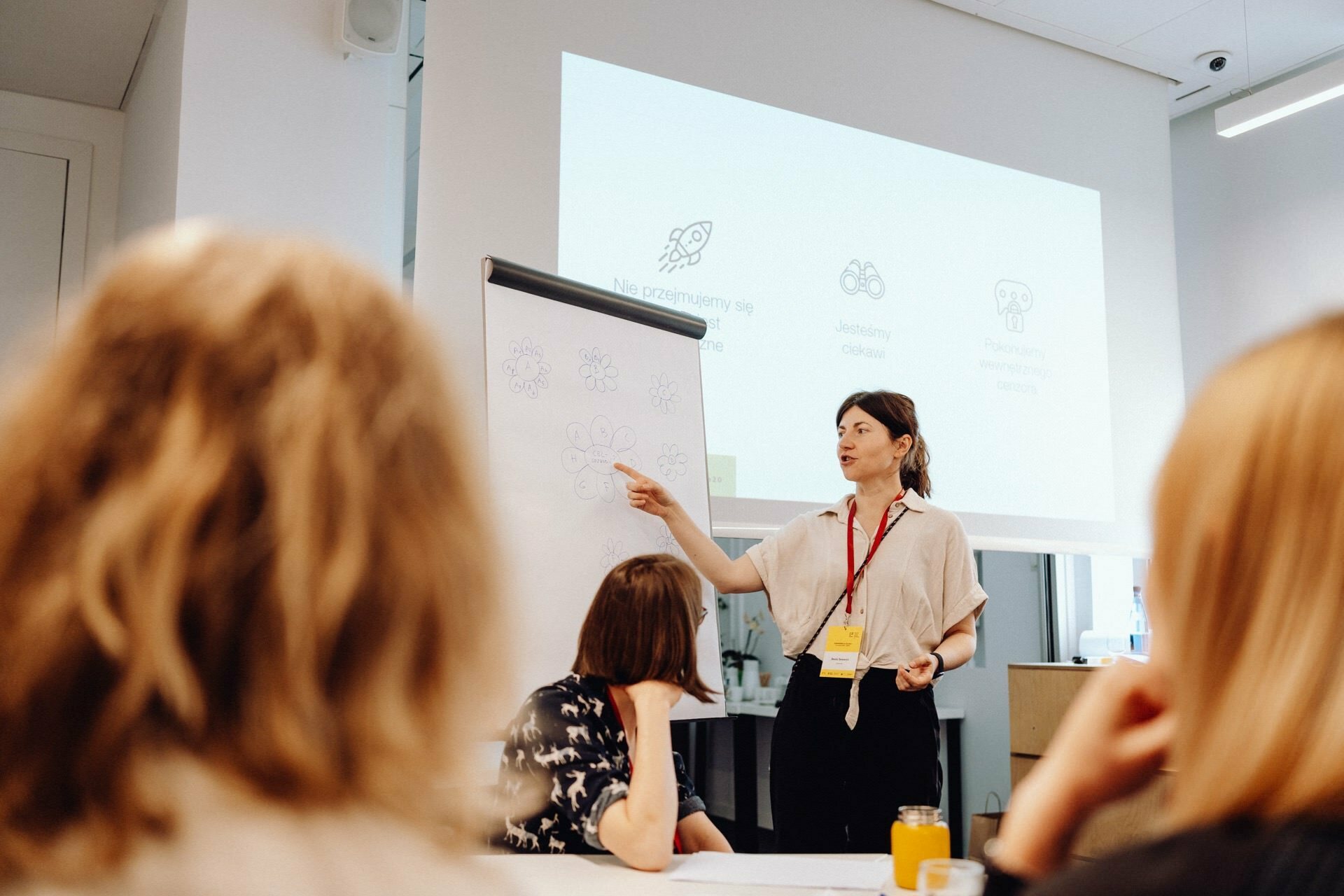 A woman presents to a group in an office. She stands next to a flipchart with several drawings on it and points to it. Behind her is a projector screen displaying icons and text. Several people sit and carefully watch her presentation while a photographer at the event captures the moment.   
