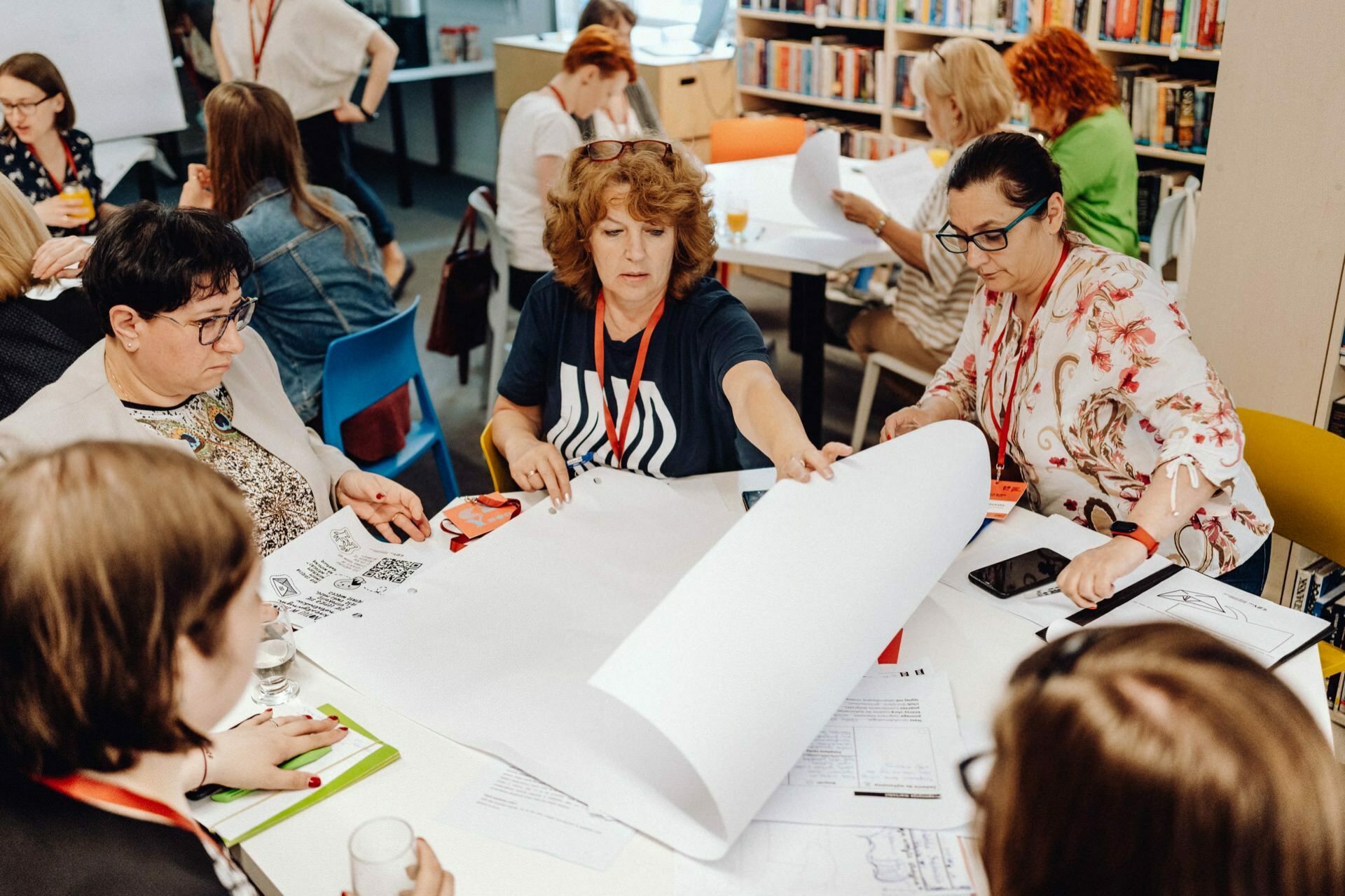 A group of people, mostly women, sit around a table in a room filled with bookshelves. They are engaged in a common activity, discussing and reviewing large sheets of paper. One woman in the middle is unfolding a large paper, while others are watching - a scene perfect for event photo coverage by any skilled event photographer Warsaw.  