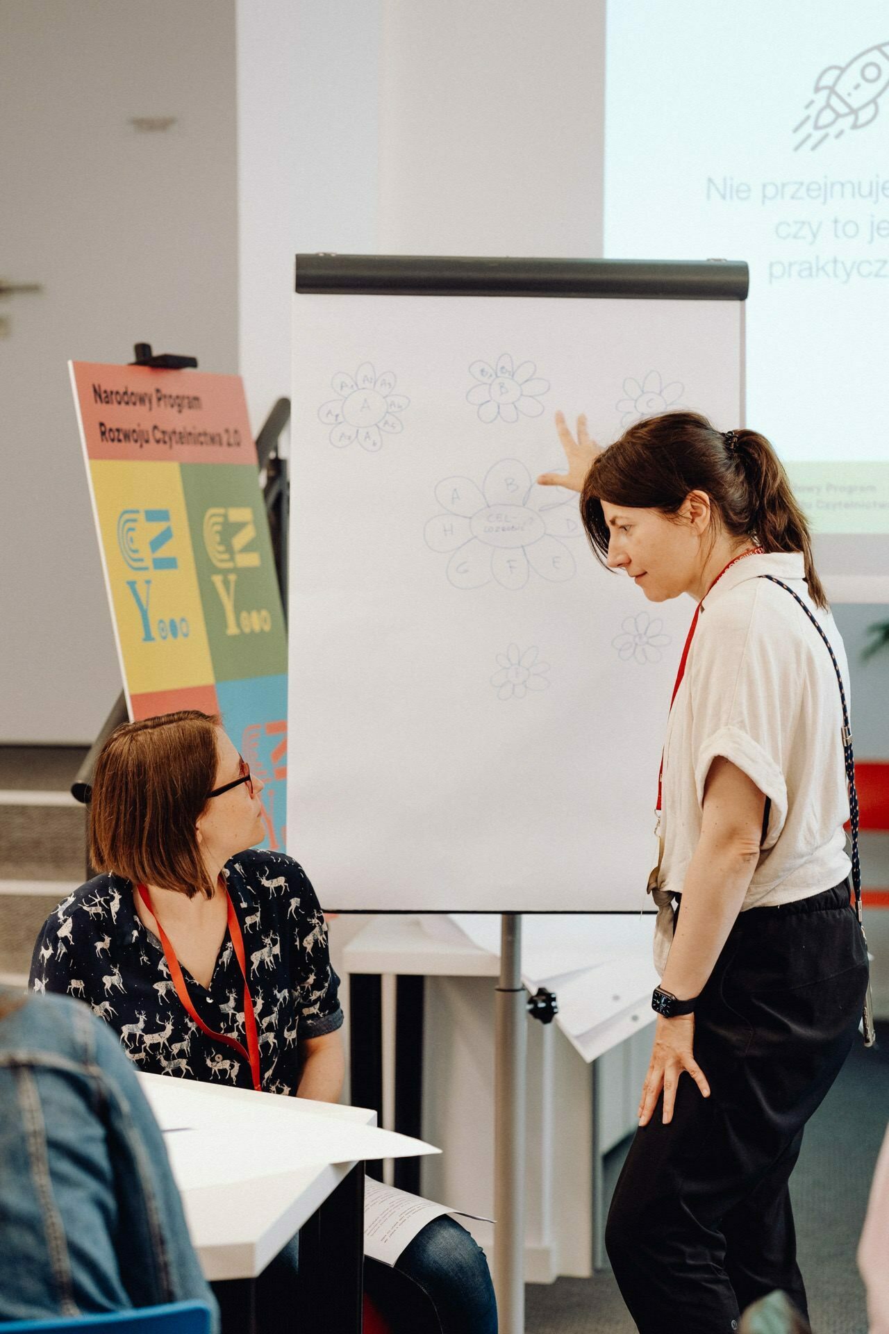 Two women are discussing at a flipchart stand, while one stands and points to a diagram on paper. The diagram consists of flower shapes and circles connected by lines. The seated woman observes. Both are wearing red lanyards. A banner is visible in the background, perfectly captured by the photographer at the event.    