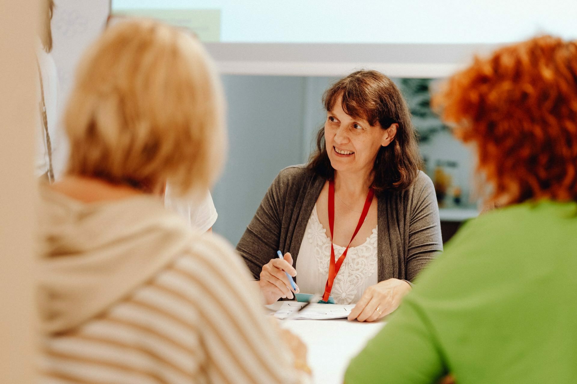 A woman with brown hair and a red leash smiles as she sits at a table, holding a pen and paper. She is engaged in a conversation with two people, one with blonde hair and the other with red curly hair, who are sitting across from her. In the background is a projection screen that captures the essence of the photo-report of the event.  