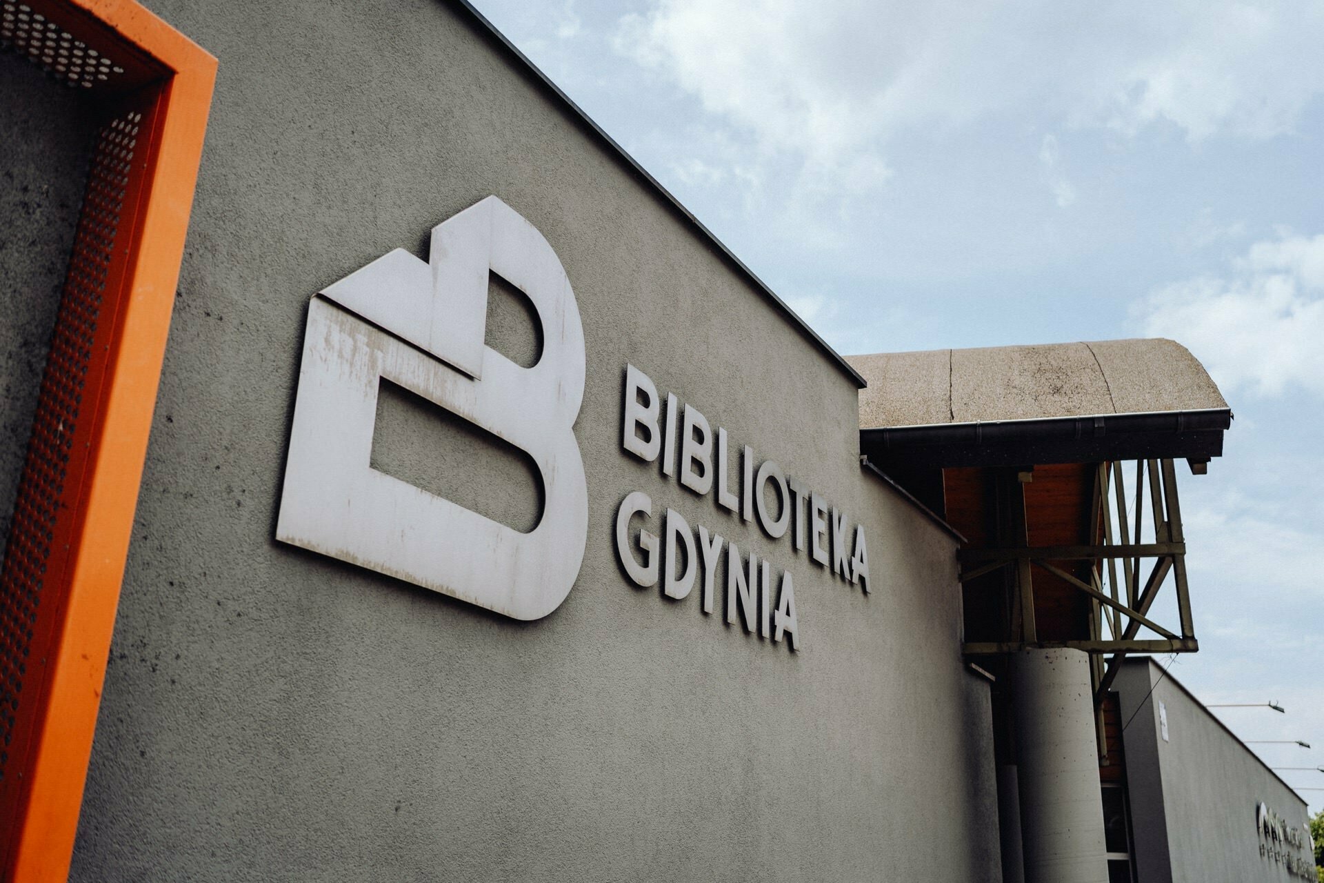 The building's wall features a large white logo and the text "Library Gdynia." The building has a modern design, characterized by clean lines and a combination of concrete and wooden elements, with a partly cloudy sky in the background. A talented photographer from Warsaw captured this moving scene.  