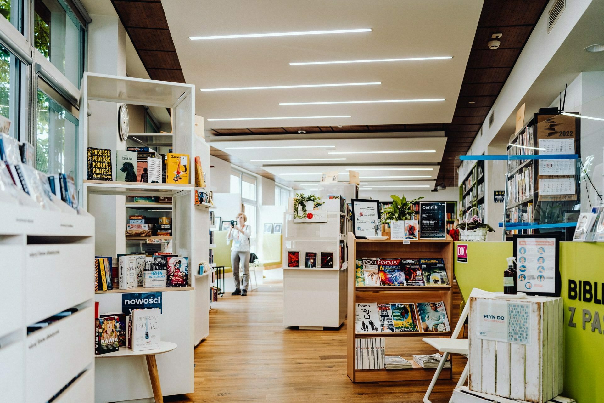 A modern library with wooden floors and bright lighting is furnished with neatly arranged shelves of books and magazines. Several people browse through the collection. Large windows on the left let in natural light, perfect for capturing the event of a photographer from Warsaw. There is a plant and informational posters at the entrance.   