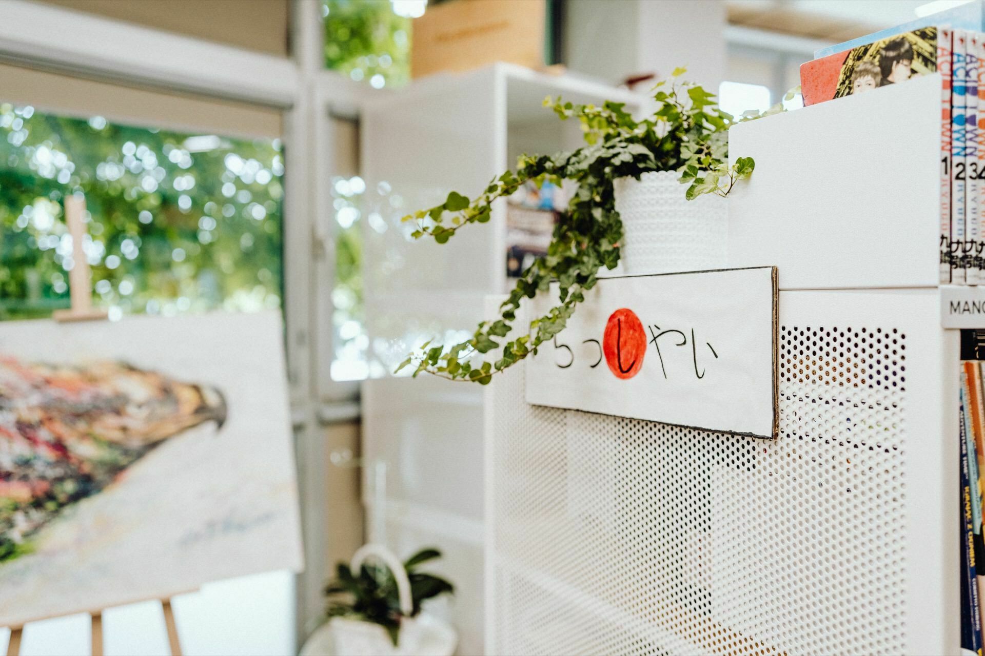 A bright room with white shelves decorated with a green plant. A sign with Japanese characters and a red circle is displayed on the shelf. In the background, a blurry painting on an easel shows an abstract scene with a variety of colors, perfectly captured by event photographer Warsaw.  