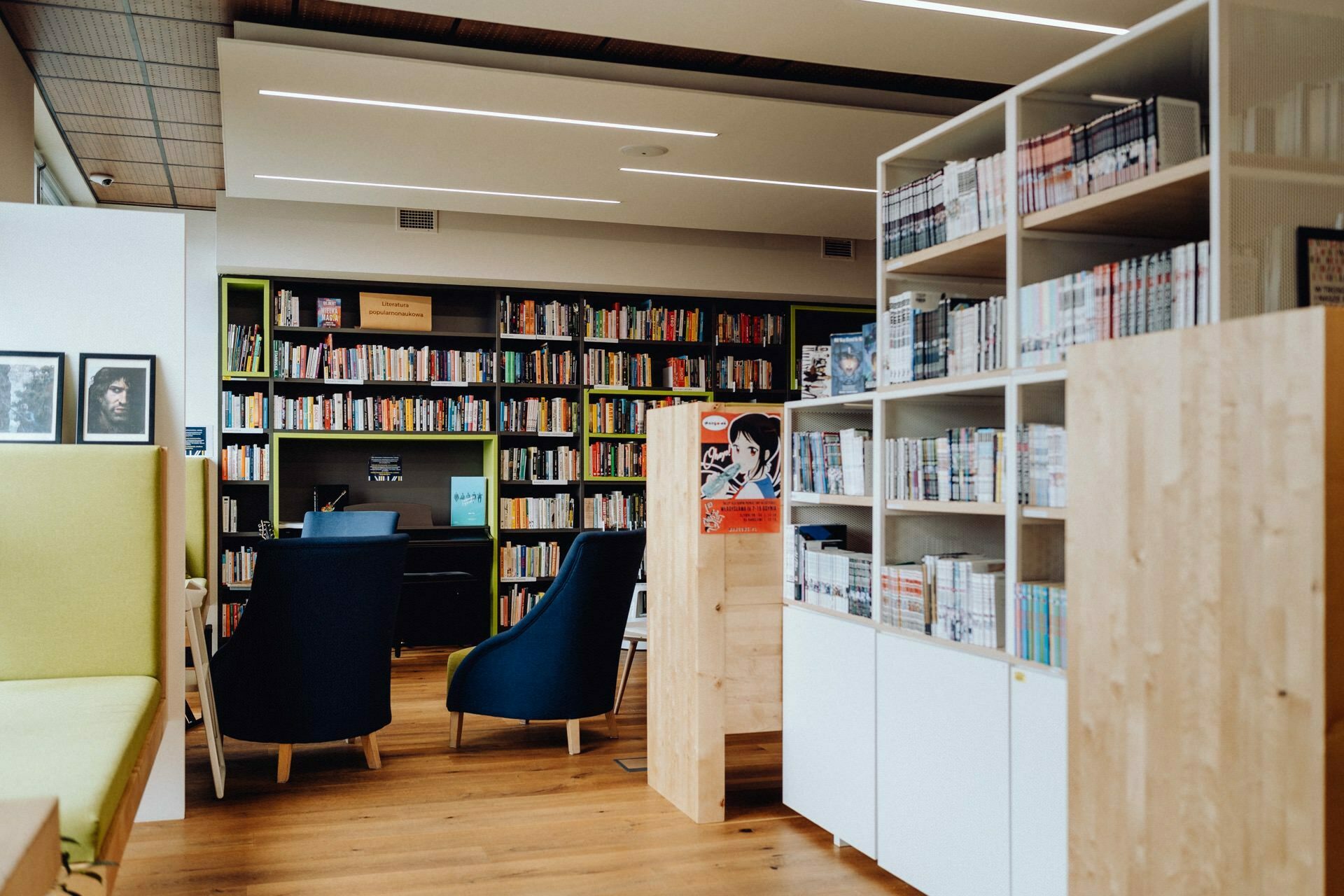 A modern library interior with wooden floors, shelves filled with books and cozy seating areas. Two blue armchairs stand next to a small table in front of a bookshelf in the background. Shelves and more books are visible in the foreground, perfect for a Warsaw event photographer to capture its charm.  