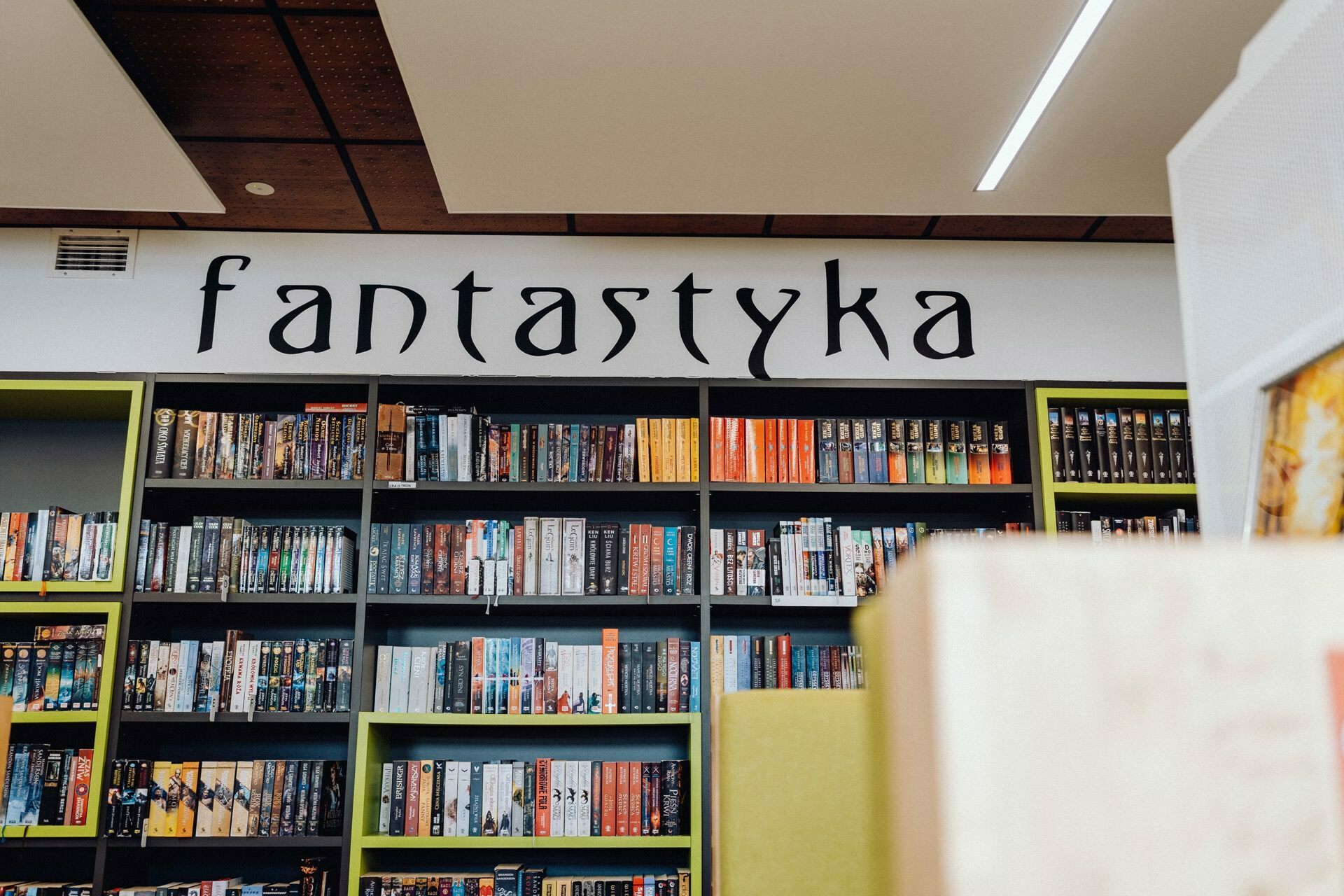 The brightly lit section of the bookstore labeled above "fantasy" with black shelves filled with numerous fantasy books. The shelves are organized with colorful book spines. In the foreground, a blurry, partial view of nearby books and clues about the photographer at the event, capturing the magical atmosphere.  