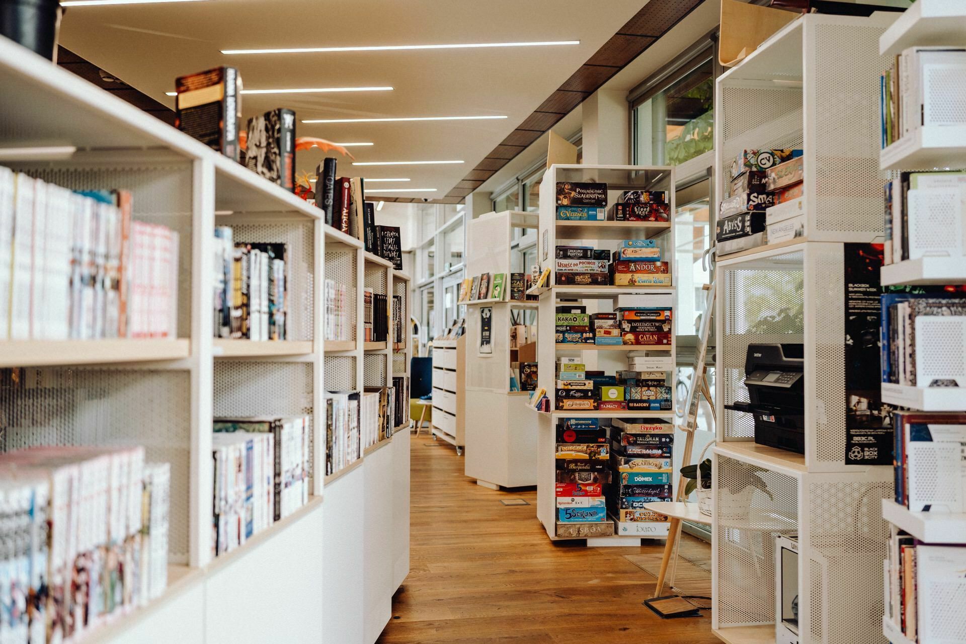 A modern library with wooden floors and white shelves filled with books and board games. The space is well lit with natural light streaming in through large windows, perfect for a Warsaw event photographer to capture its charm. The shelves are neatly organized, creating a calm and inviting atmosphere.  