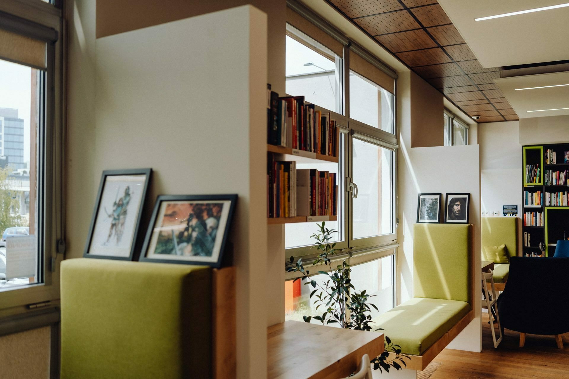 A cozy reading nook with green padded benches, framed pictures and a bookshelf filled with books. Large windows let in natural light, creating a warm and inviting atmosphere, perfect for a photography party. Potted plants and modern decor complete the look.  