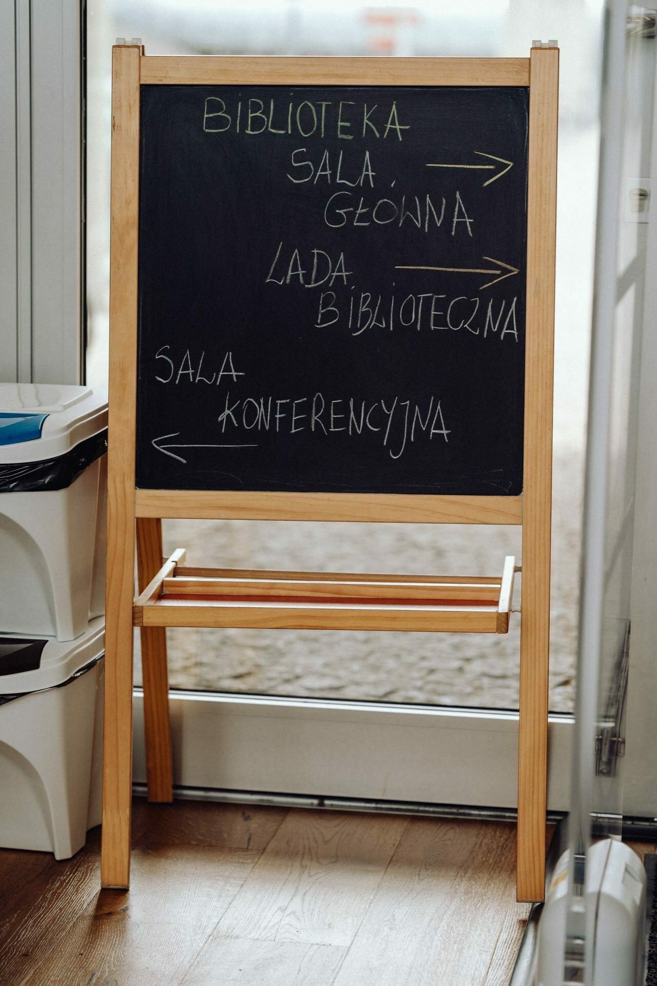 A blackboard with wooden legs stands in a room. It contains handwritten directions in Polish: "BIBLIOTEKA" at the top, with arrows pointing to "MAIN ROOM," "LIBRARY ROOM" and "CONFERENCE ROOM." A photographer from Warsaw captured this scene, next to which various storage containers are partially visible  