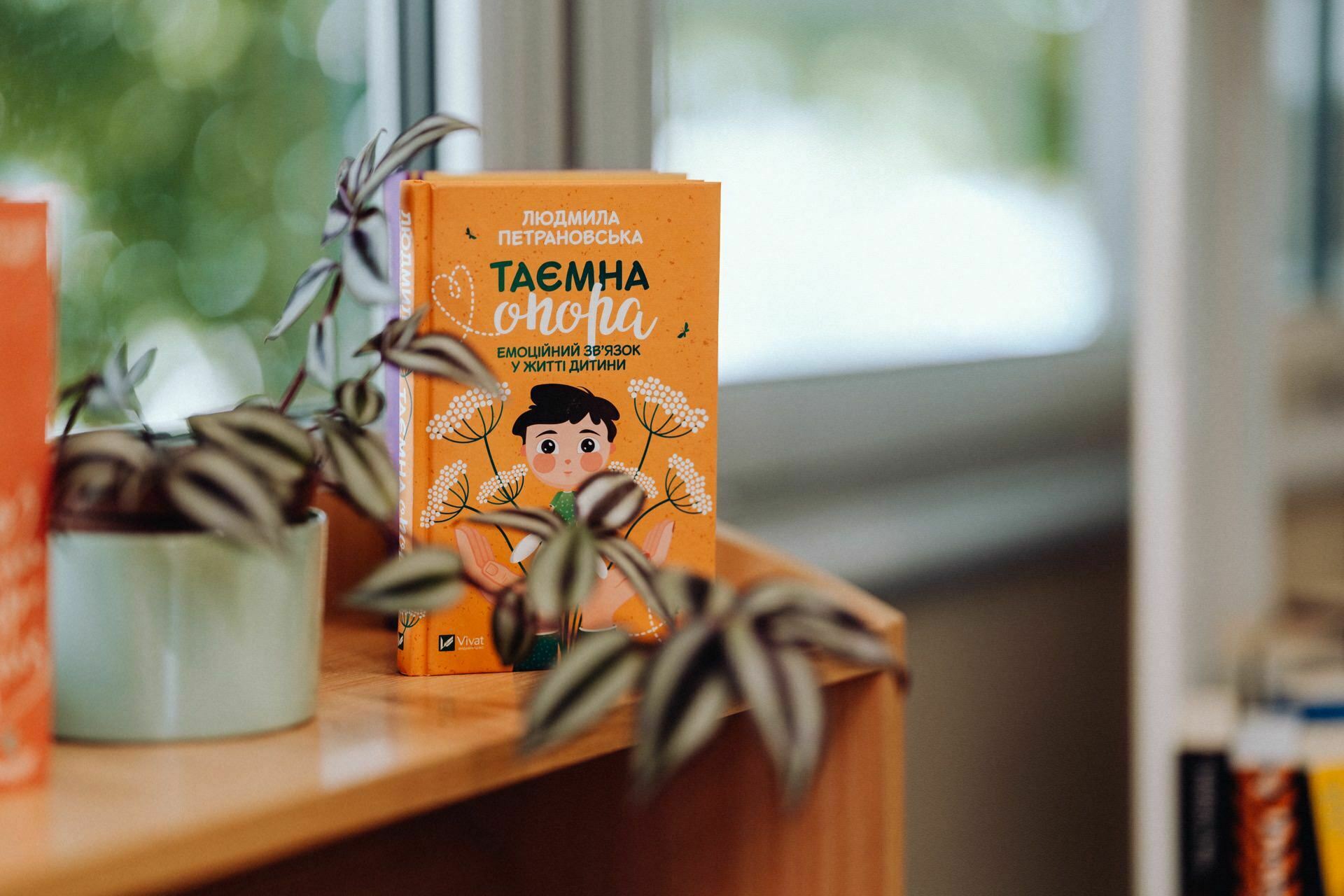 An orange booklet with an illustration of a child holding a green plant is placed on a wooden surface next to a potted plant, reminiscent of an event photo. The text on the book's cover is written in a non-Latin alphabet, and a window with blurred outdoor scenery is visible in the background. 