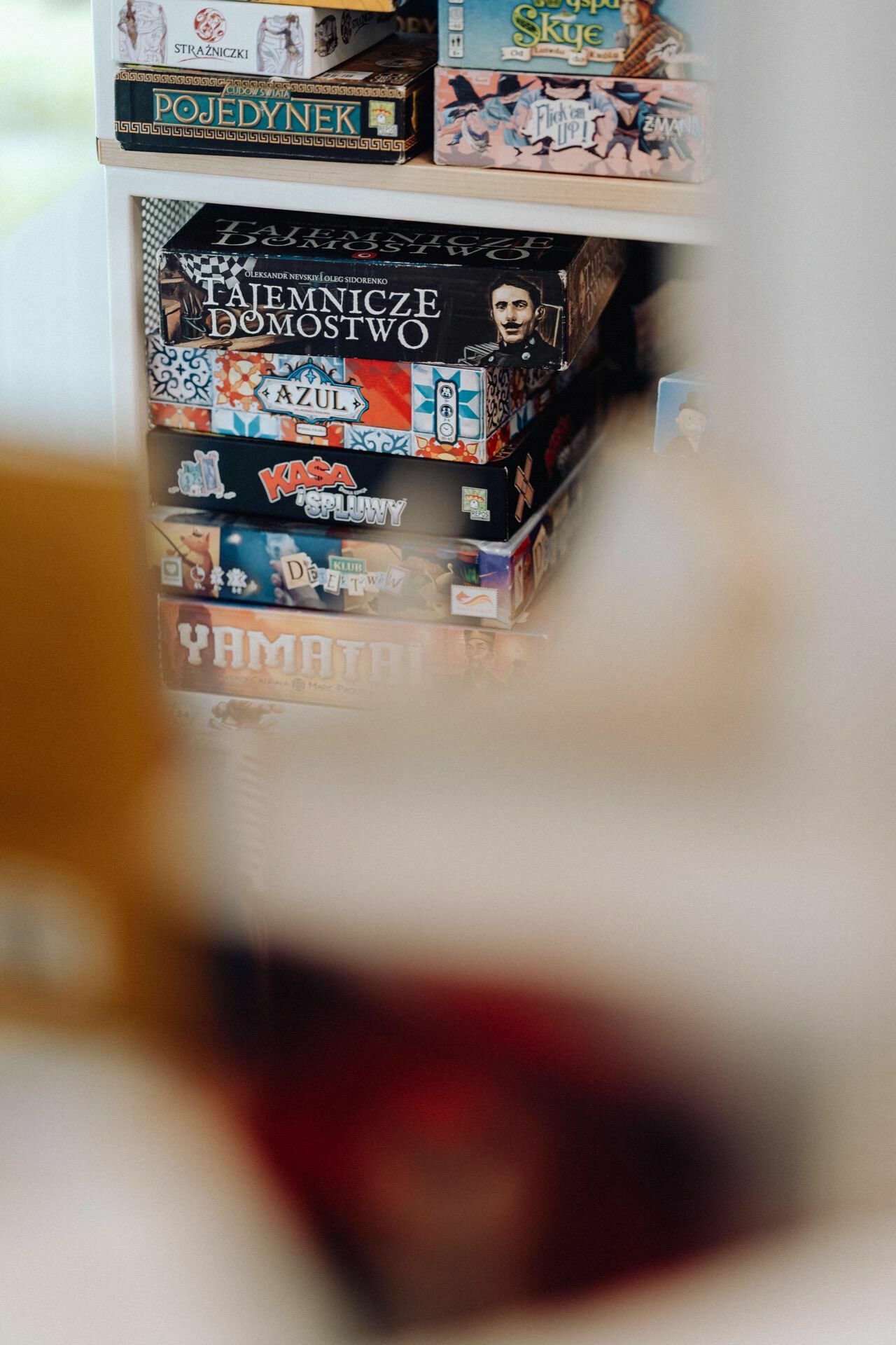 A stack of colorful board games is stacked on a shelf. Visible titles include "Secrets of the Homestead," "Azul," "Karuba," "Yamatai," and others in various languages, reminiscent of a vibrant photo-report of the event. Part of the image is out of focus, resulting in a slight blurring around the edges.   