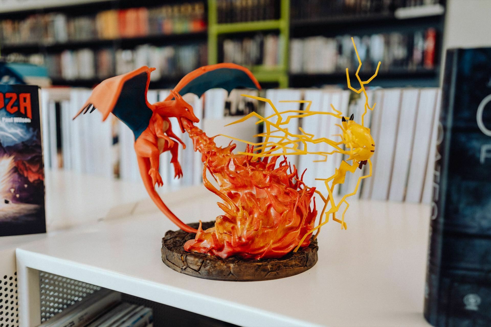 The detailed white table figurine depicts two fighting dragon-like creatures. One orange creature lets out a stream of fire, while the other yellow creature responds with electric sparks. In the background is a bookcase filled with various books, beautifully captured by a photographer from Warsaw.  