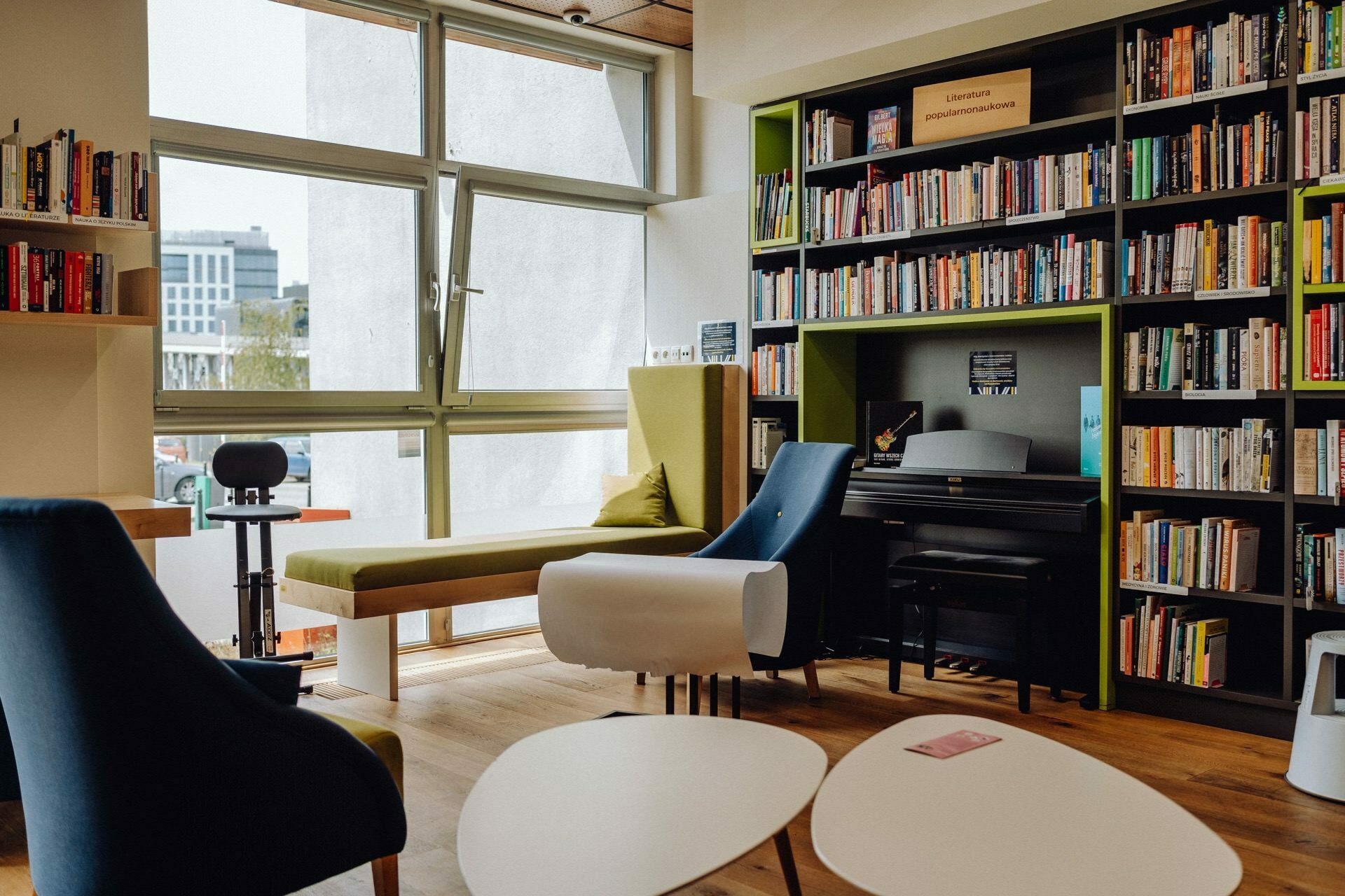 The cozy reading corner is equipped with comfortable chairs, a folding bench by the window and a small table. The walls are decorated with shelves filled with books, and a piano stands in the corner. Natural light streams in through the large windows, creating a warm atmosphere ideal for capturing photo coverage of the event.  