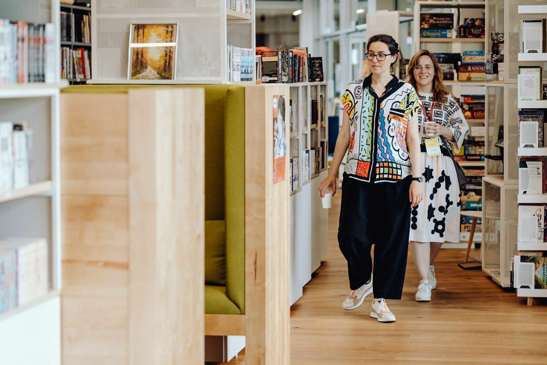 Two women walk through a library, both wearing colorful patterned blouses. The woman in front has dark hair tied back and glasses, the woman behind her has long, light hair. They smile as they pass shelves of books and magazines that perfectly capture the events the photographer na.  