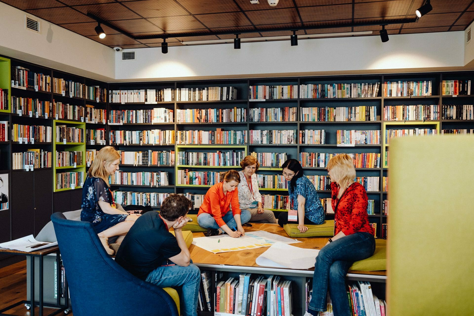 A group of six people collaborate at a table in the library. The table is cluttered with papers and books, perfect for a photo-op of the event. Bookshelves filled with colorful books line the background, creating a cozy and learning atmosphere.  