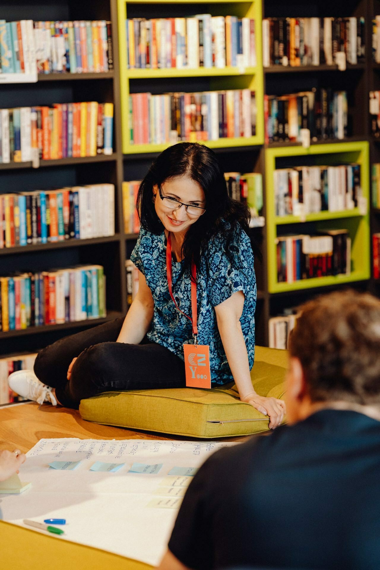 A woman with glasses sits on a cushion in the library and is having a discussion with the person in front of her. She has a lanyard around her neck and papers spread out in front of her. In the background are colorful shelves filled with books, captured by Event Photographer Warsaw.  