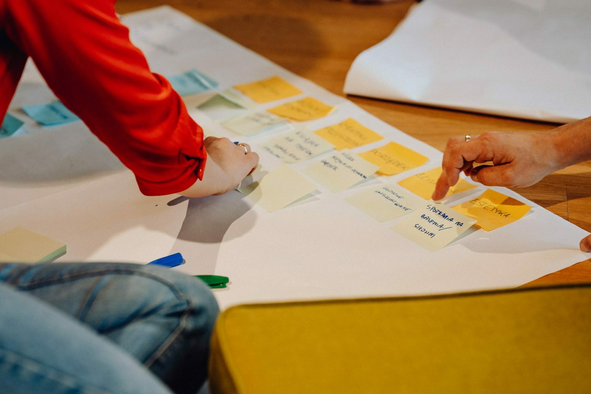 Two people arrange colored post-it notes on a large sheet of white paper spread out on a wooden surface. One person in a red shirt is kneeling, the other person's hand is visible. Blue and green markers are scattered nearby, perfectly captured by a photographer from Warsaw who specializes in event photography.  