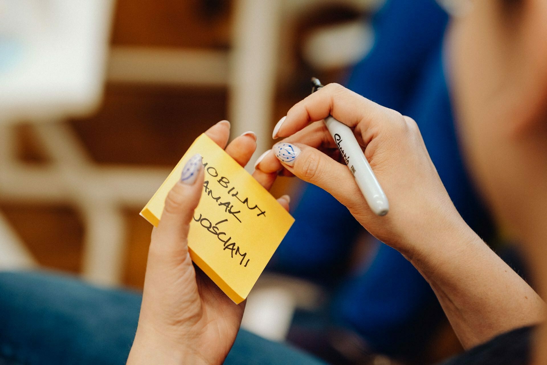 A person with light blue nail polish and decorative nail ornaments holds a yellow sticky note and writes on it with a black Sharpie marker. On the note are words written in black ink. The background is blurry and indistinct, reminiscent of an event photo by a photographer for an event in Warsaw.  