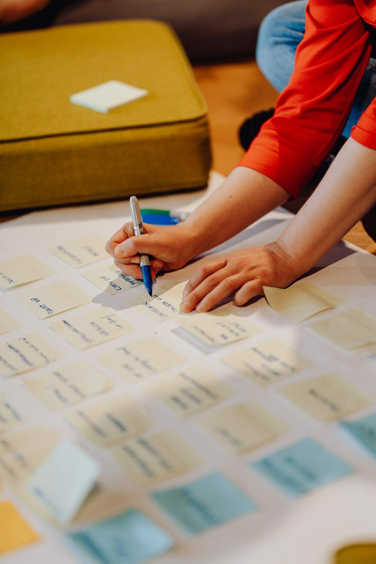 A person wearing an orange shirt leans over the table and writes on a sticky note with a blue pen. The table is covered with numerous yellow, blue and green sticky notes, some with visible text. In the background is a green pillow, perfect for any photo event.  