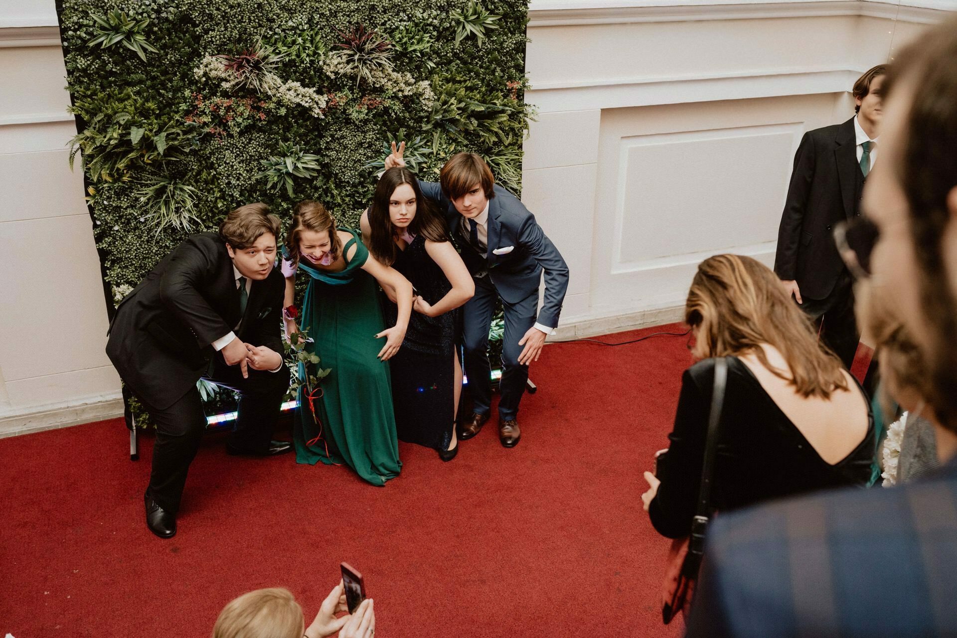 A group of four young adults, dressed formally, playfully pose for a photo against a background of green leaves. Standing on a red carpet, they make various gestures and smile. Around them, other participants can be seen capturing moments on their devices, as if in a bustling report from a prom.  