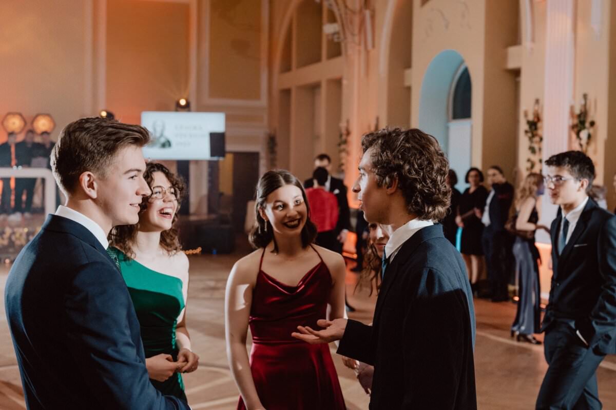 Grupa elegancko ubranych młodych dorosłych prowadzi ożywioną rozmowę podczas oficjalnego wydarzenia w wielkiej, ciepło oświetlonej sali. Niektórzy uśmiechają się, podczas gdy ktoś mówi z ożywieniem. Inni w tle mieszają się i cieszą atmosferą przypominającą reportaż ze studniówki.