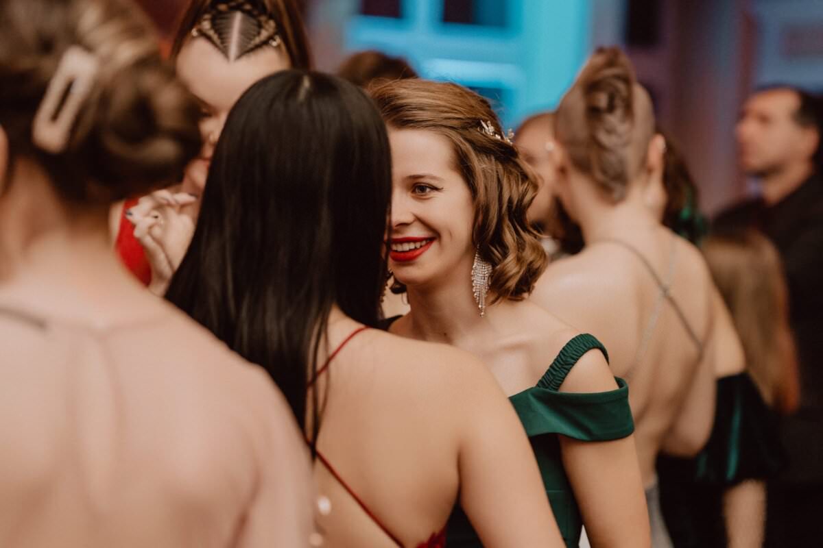 Grupa kobiet ubranych w elegancki, formalny strój rozmawia blisko podczas imprezy towarzyskiej odbywającej się pod dachem. Jedna kobieta z krótkimi, stylizowanymi włosami i zieloną sukienką odsłaniającą ramiona uśmiecha się promiennie, stojąc twarzą w twarz z kobietą w czerwonej sukience i długimi czarnymi włosami. Przypomina to reportaż ze studniówki utrwalający cenne wspomnienia.