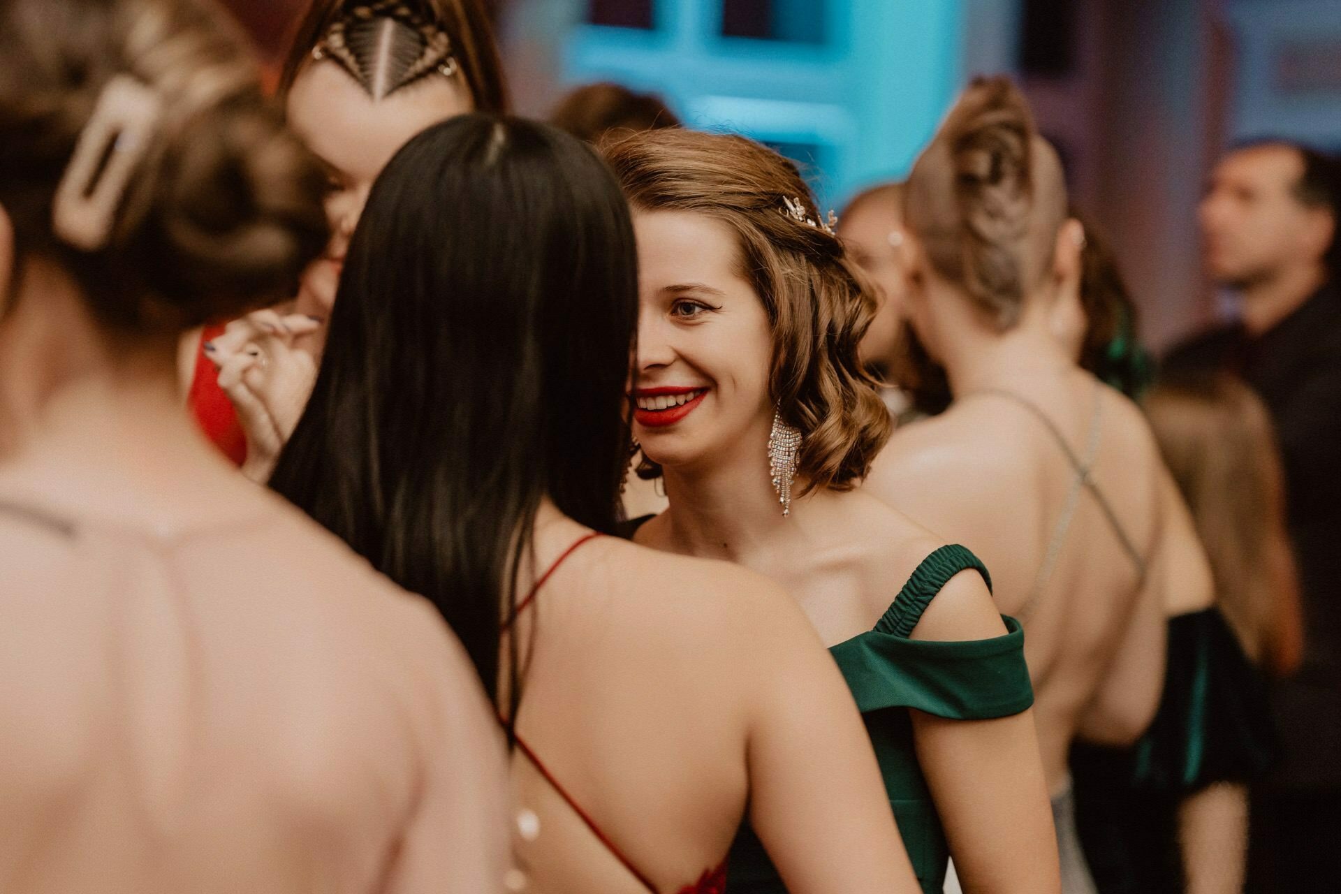 A group of women dressed in elegant, formal attire chat closely during a social event held under a roof. One woman with short, stylized hair and a green dress revealing her shoulders smiles radiantly while facing a woman in a red dress and long black hair. It is reminiscent of a reportage from a prom capturing precious memories.  