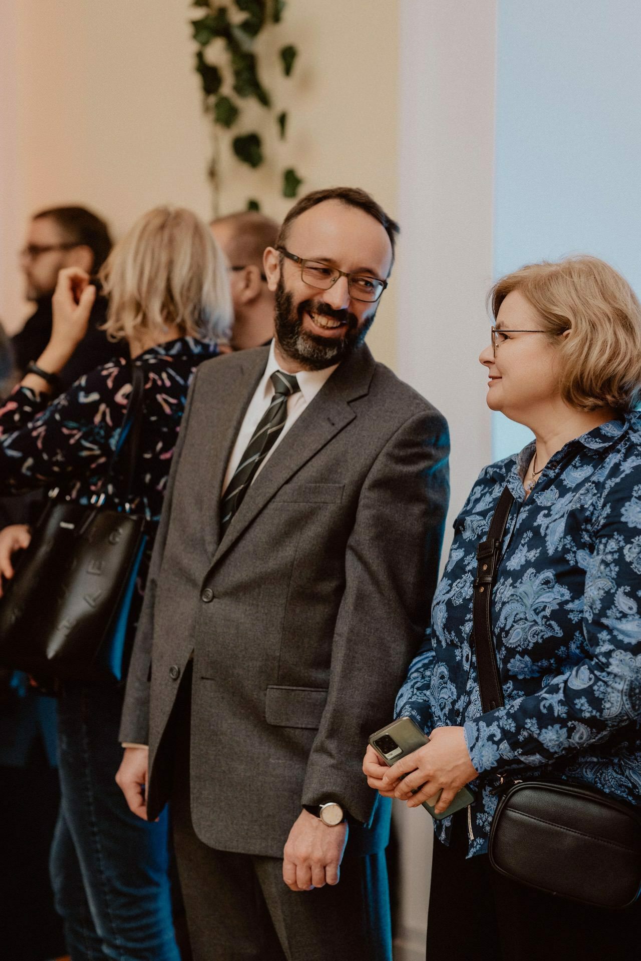 A man in a gray suit and glasses smiles while talking to a woman in a blue floral blouse during a report from a prom. The woman holding a phone and wearing glasses looks busy talking. Several other people are talking in the background, and the walls are decorated with greenery.  