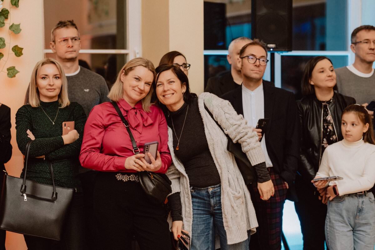 Grupa ludzi, w tym mężczyźni i kobiety, stoi razem w pomieszczeniu, niektórzy z uśmiechami i trzymają telefony. Dwie kobiety pośrodku pochylają się i uśmiechają do kamery. Młoda dziewczyna w białym swetrze stoi po prawej stronie, trzyma jakiś przedmiot i patrzy w bok. Wygląda jak scena z reportażu ze studniówki.