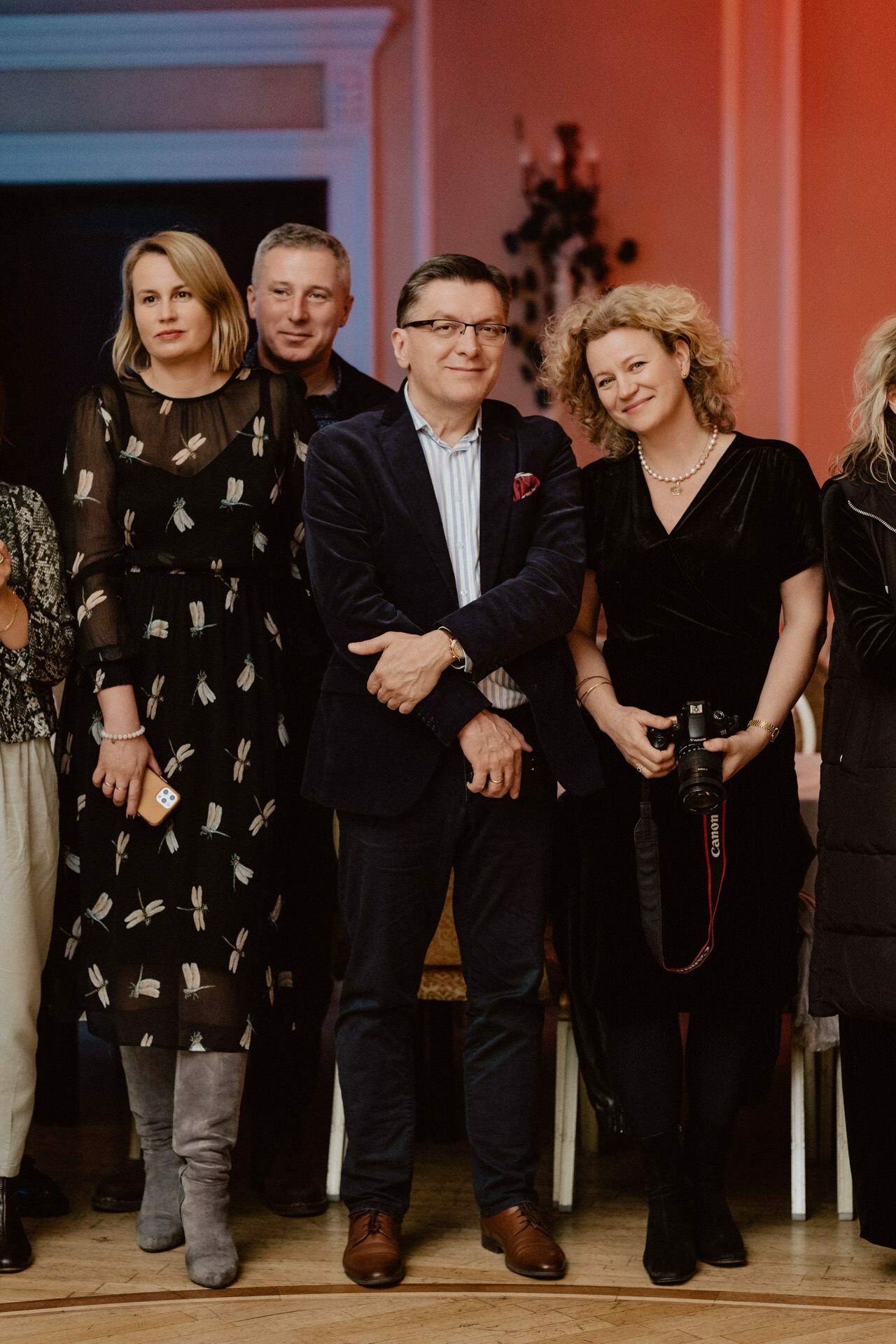 A group of five adults, three women and two men, stand in a room dressed in formal attire. One of the women is holding a camera and capturing a moment as part of a reportage from a prom. The background features warm orange lighting and dark wood finishes, creating an elegant atmosphere.  