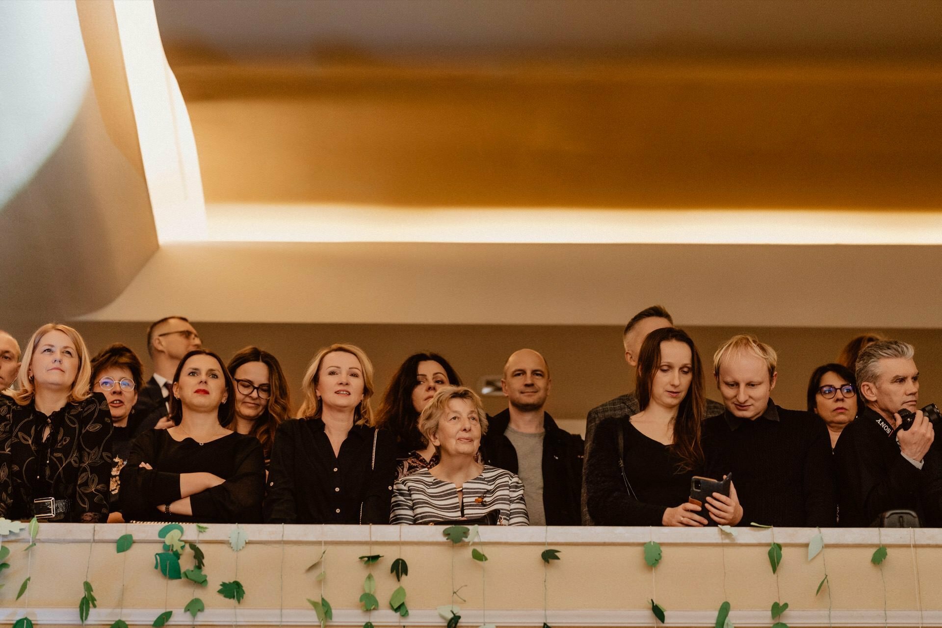 A group of people standing on a balcony and looking ahead. The balcony is decorated with green leaves hanging from the railing, evoking a sense of celebration. The setting appears to be in a room with warm lighting. Individual people show a variety of facial expressions, from attentiveness to mild curiosity, like a reportage from a prom.   