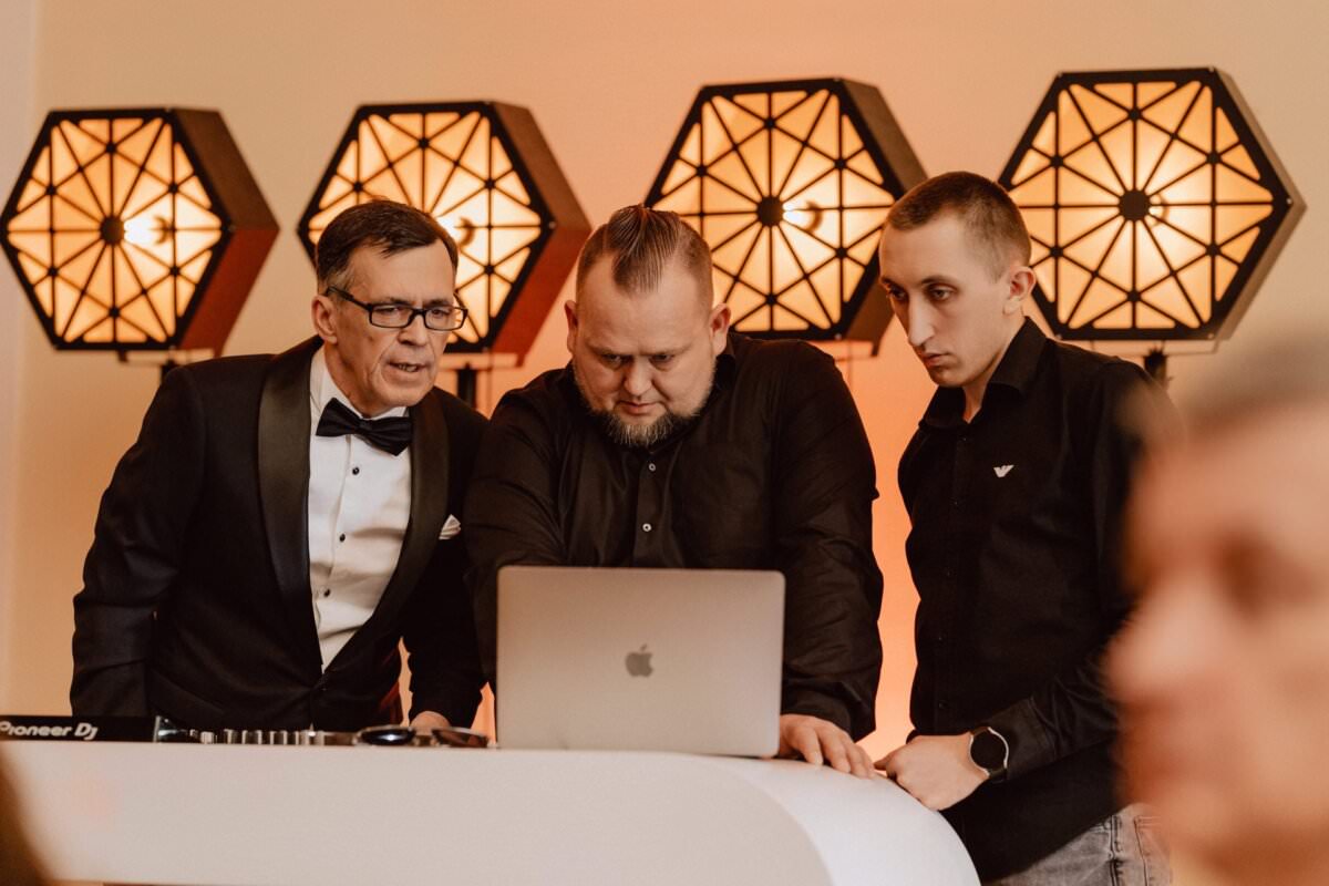 Trzej mężczyźni stoją za białym biurkiem i uważnie patrzą na laptopa. Mężczyzna po lewej stronie ma na sobie smoking, mężczyzna pośrodku ma fryzurę irokezową i jest całkowicie czarny, a mężczyzna po prawej stronie ma na sobie czarną koszulę. W tle migoczą dekoracyjne światła, jakby uchwycone w reportażu ze studniówki.